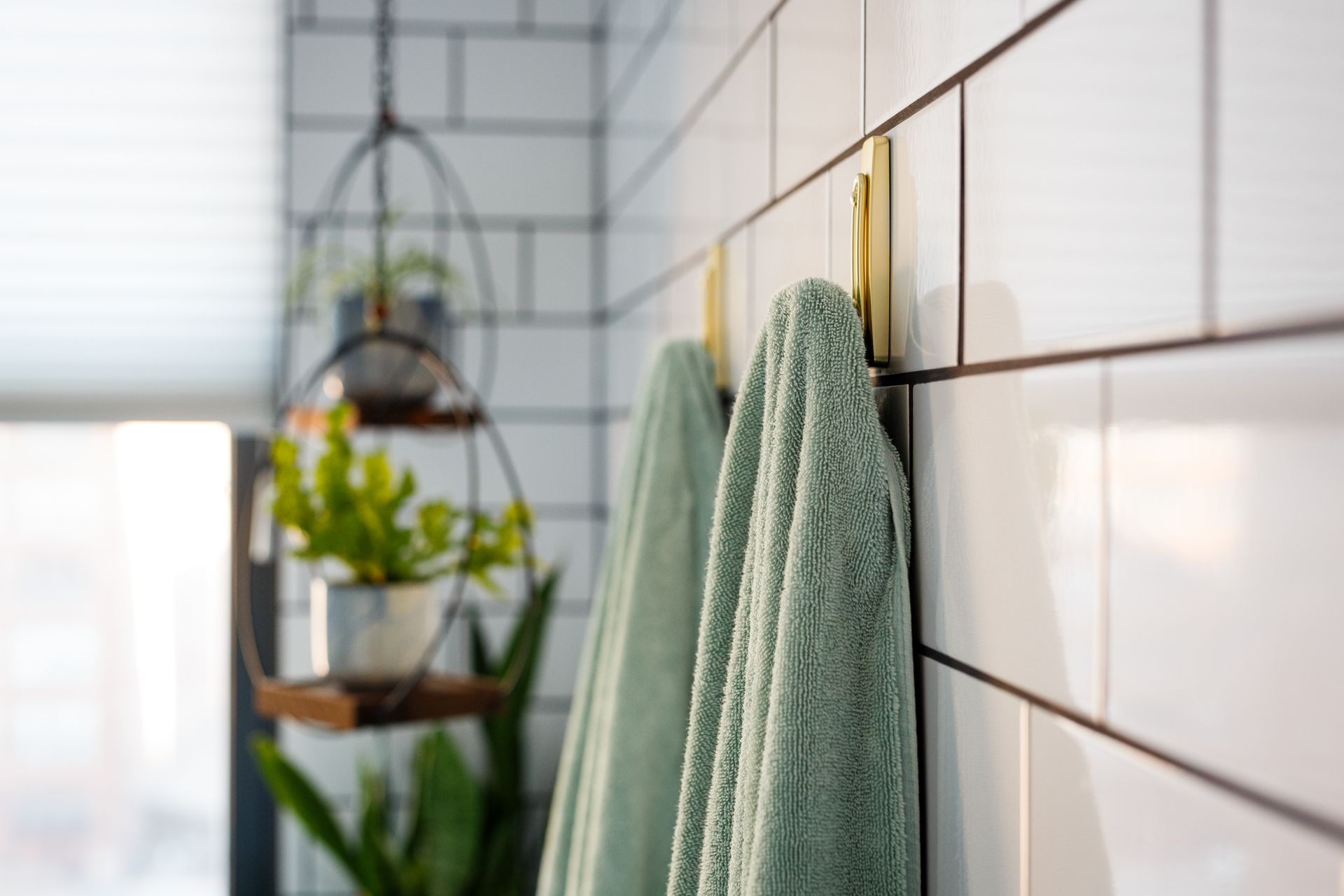 Two green towels hanging on gold hooks on white tiled wall. Shelf with plants in background.