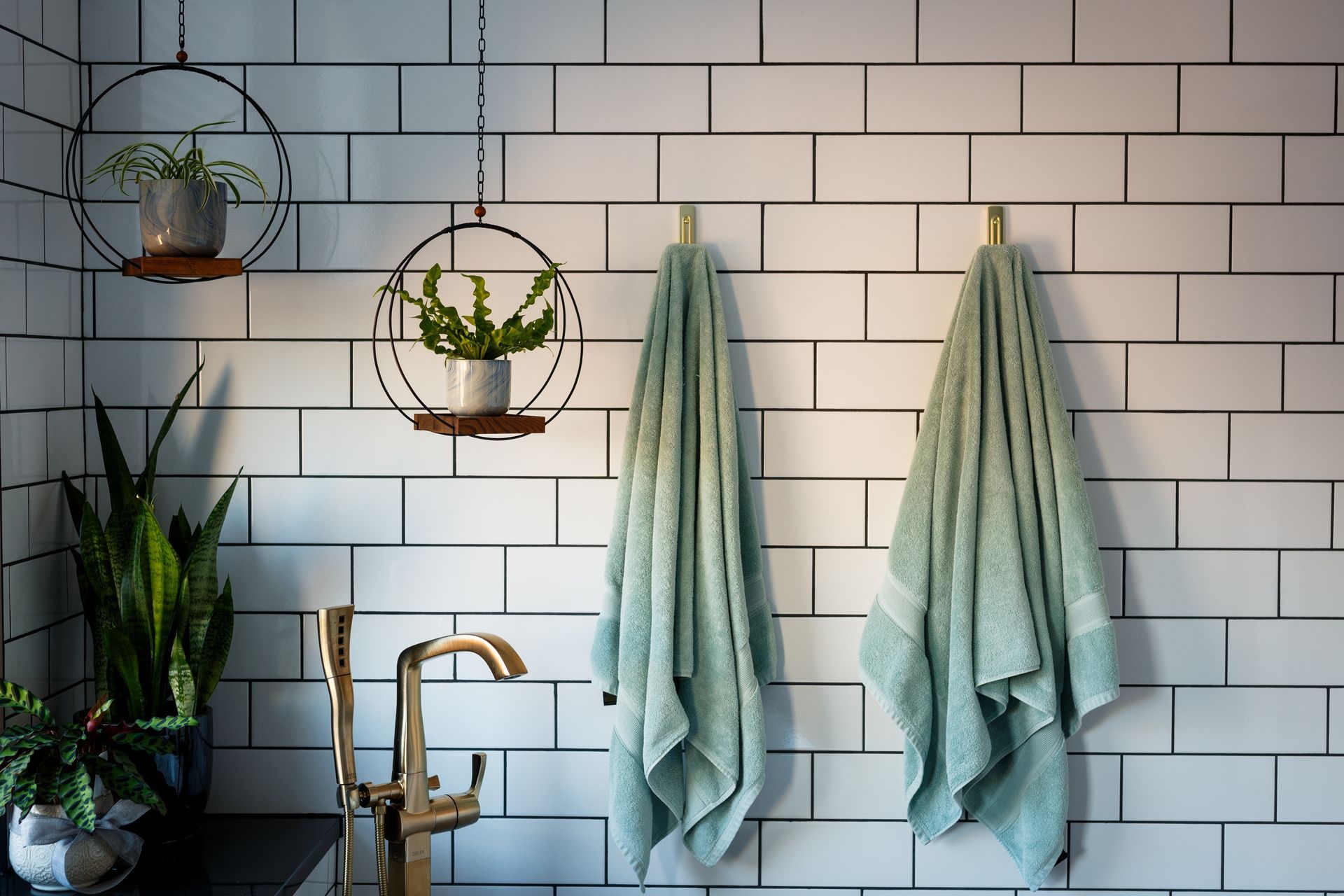 Bathroom with white subway tile, hanging towels, plants, and gold faucet.