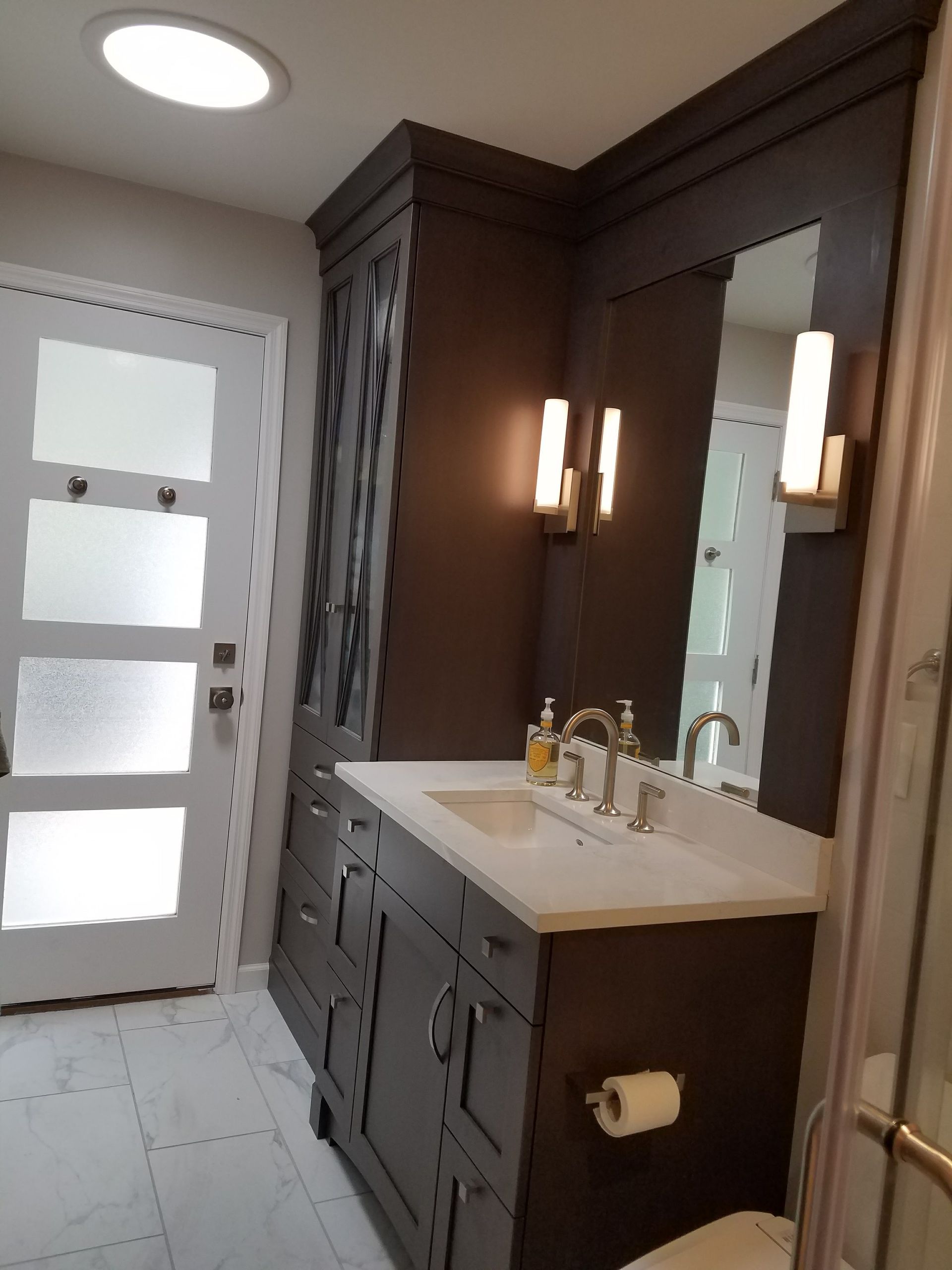 Bathroom with dark brown vanity, white countertop, sconces, frosted door, and skylight.