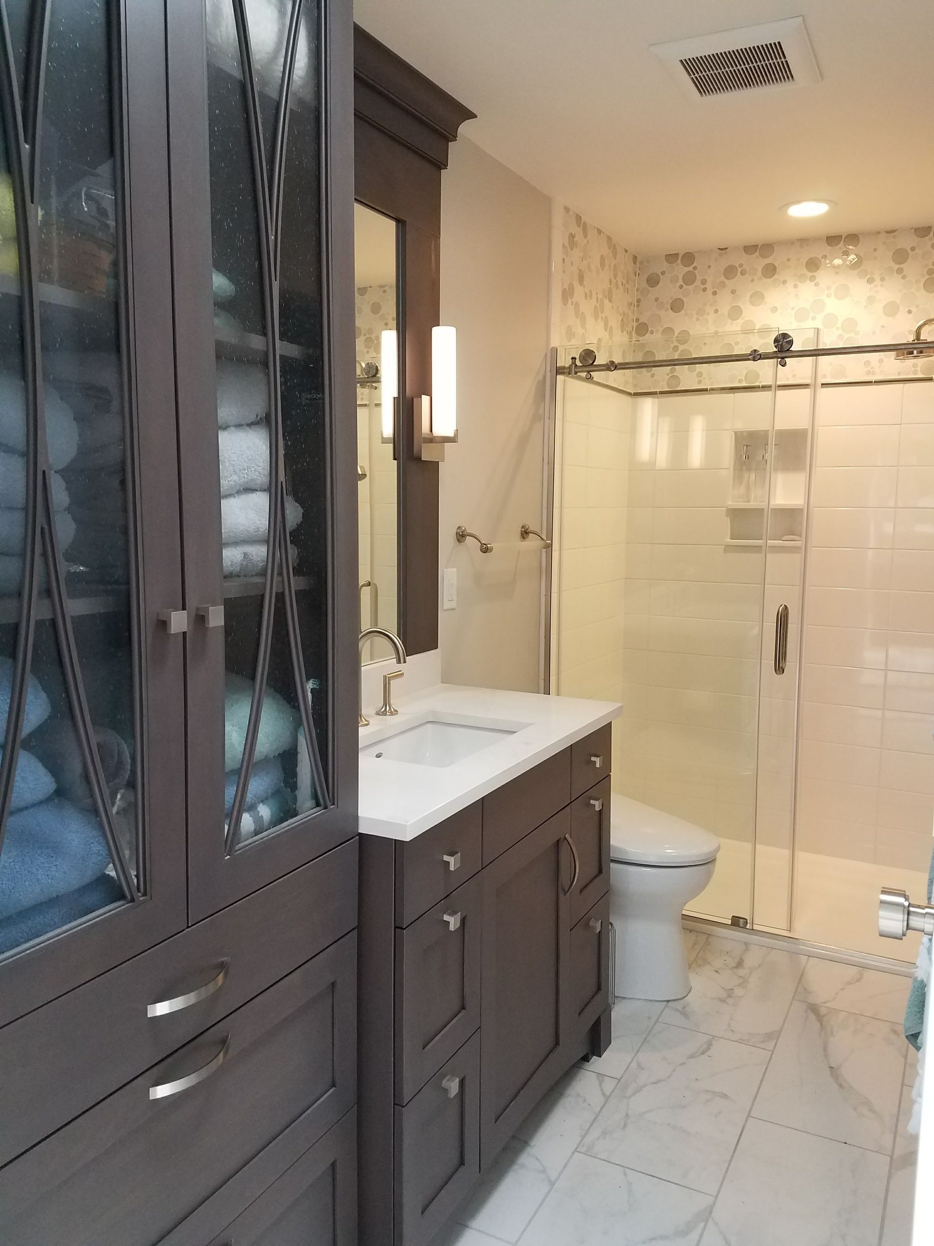 Bathroom with dark brown cabinetry, glass-enclosed shower, marble-look floor, and towels in a cabinet.