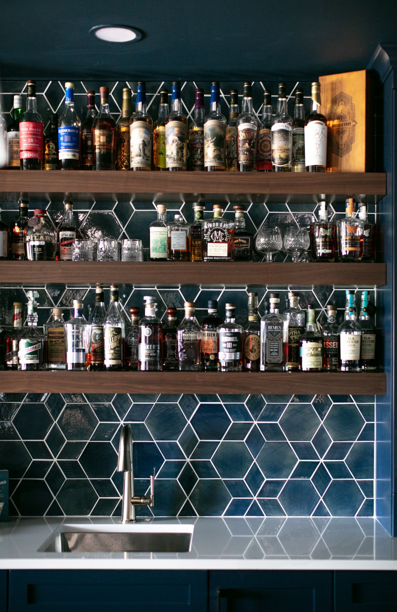 Bar shelves filled with liquor bottles and glassware, above a sink and blue geometric tile backsplash.