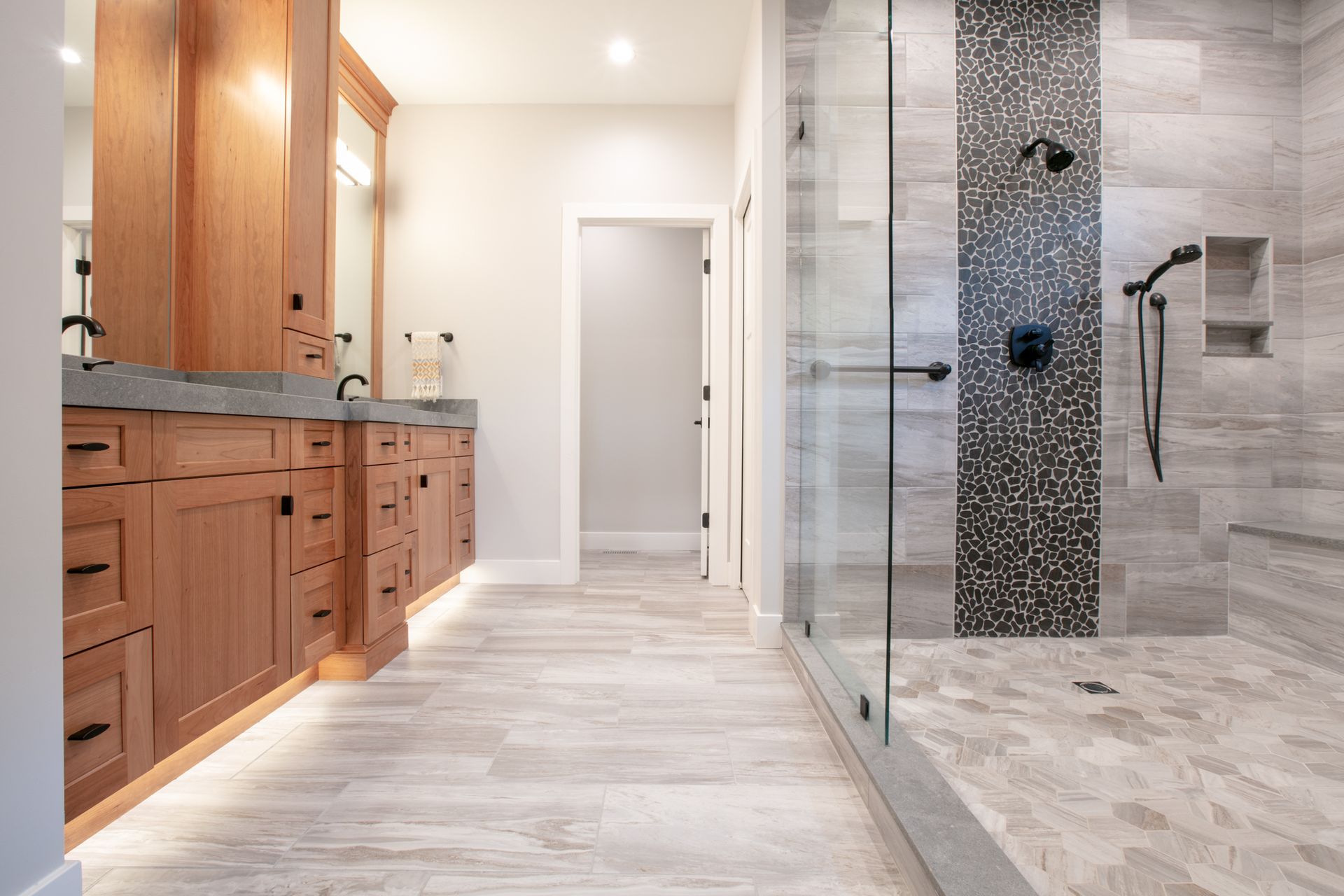 Modern bathroom with wooden vanity, gray tile floor, glass shower, and black fixtures.