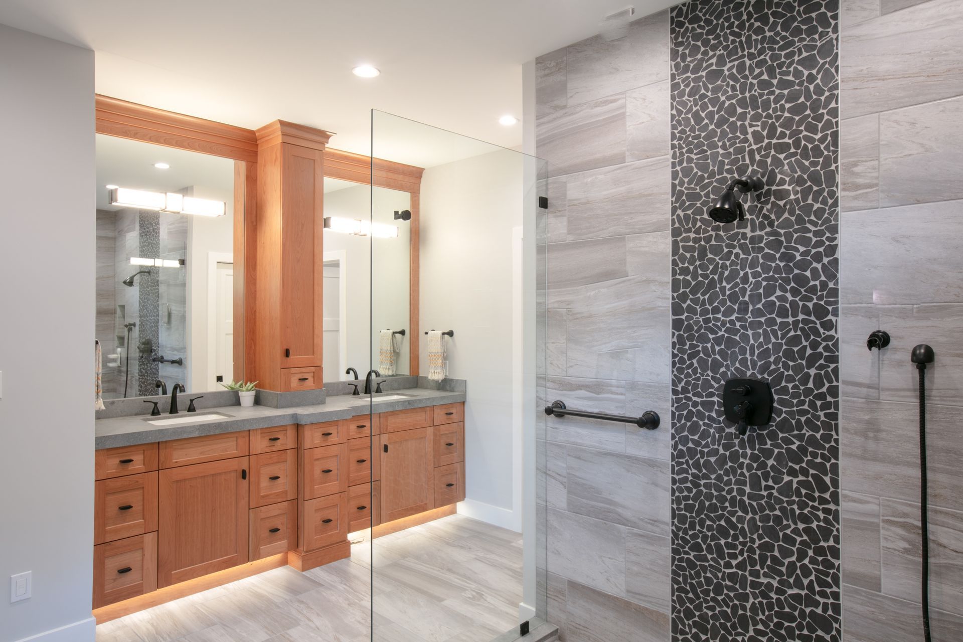 Bathroom with double vanity, large shower, gray tile, and wooden cabinets.