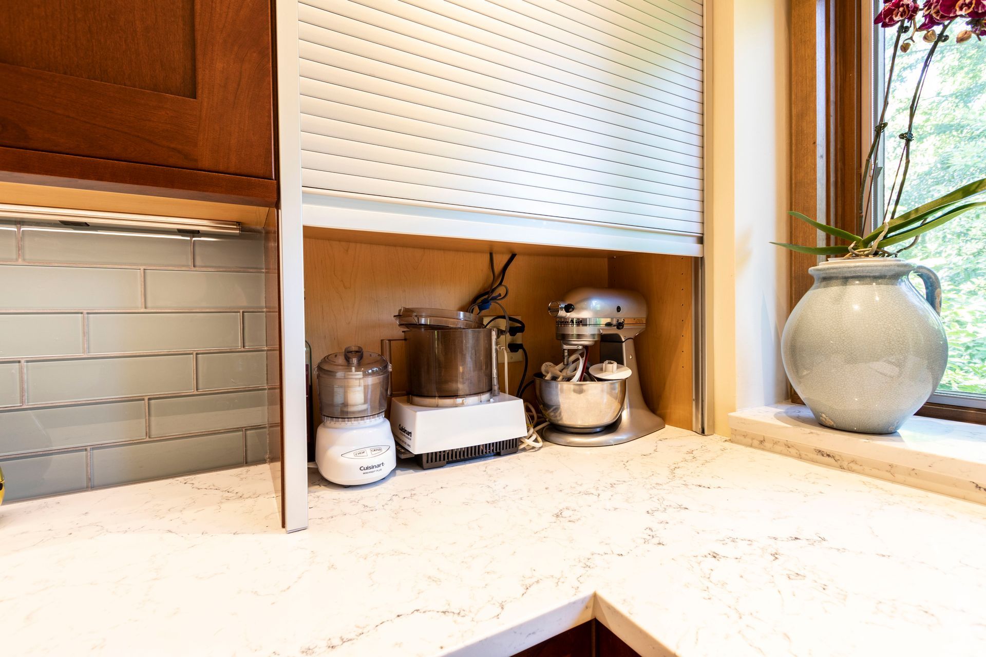Kitchen countertop with appliances inside, partially covered by a retractable white door next to a window with an orchid.