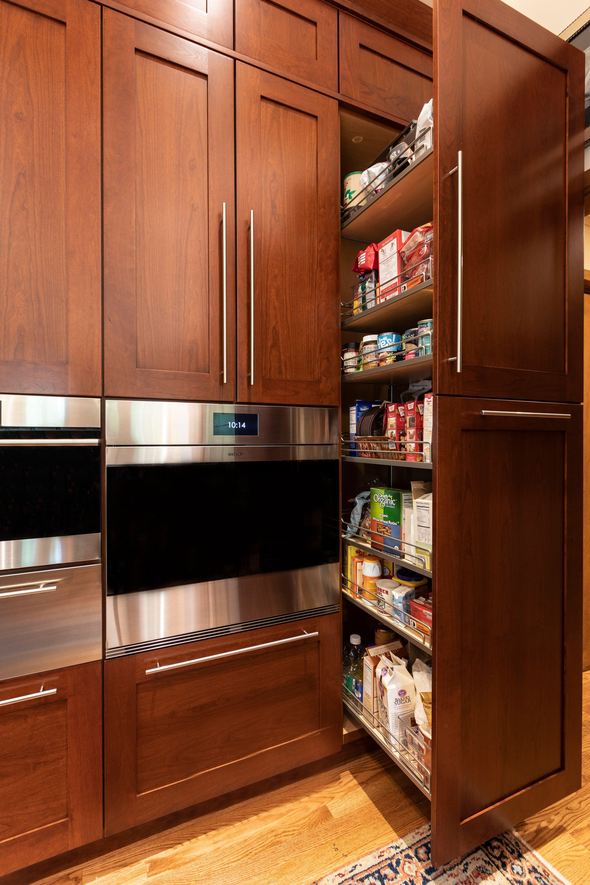 Tall, dark wood kitchen pantry cabinet with pull-out shelves, next to a stainless steel oven.