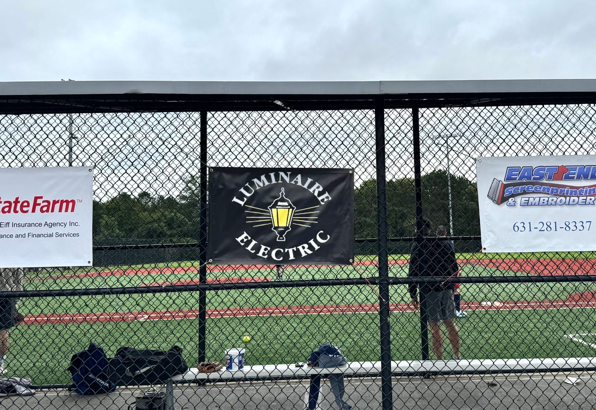 Banner reading “Illuminate Electric” on a chain-link fence beside a sports field.