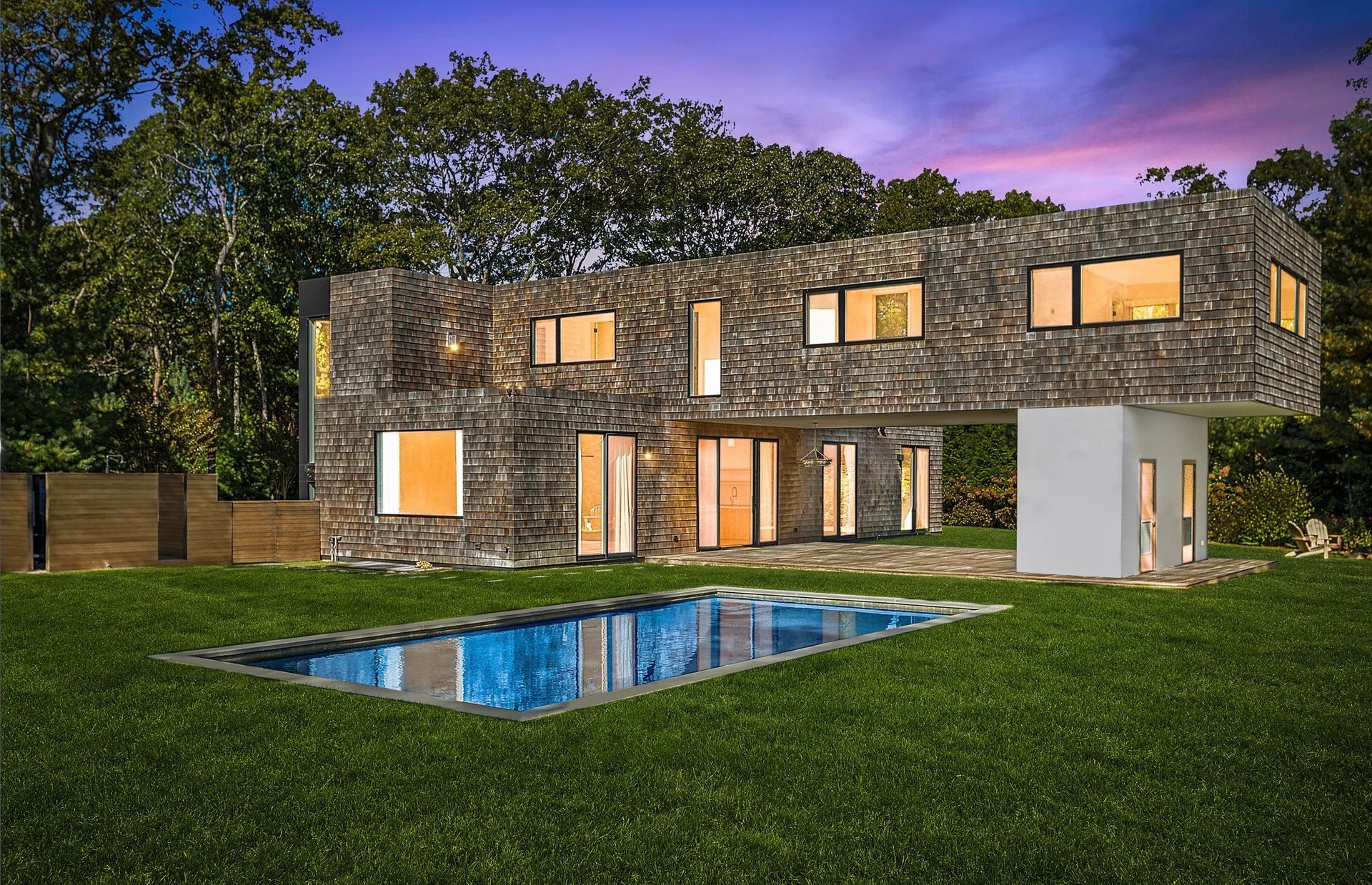 Modern two-story stone house with lit windows, a pool, and green lawn at dusk