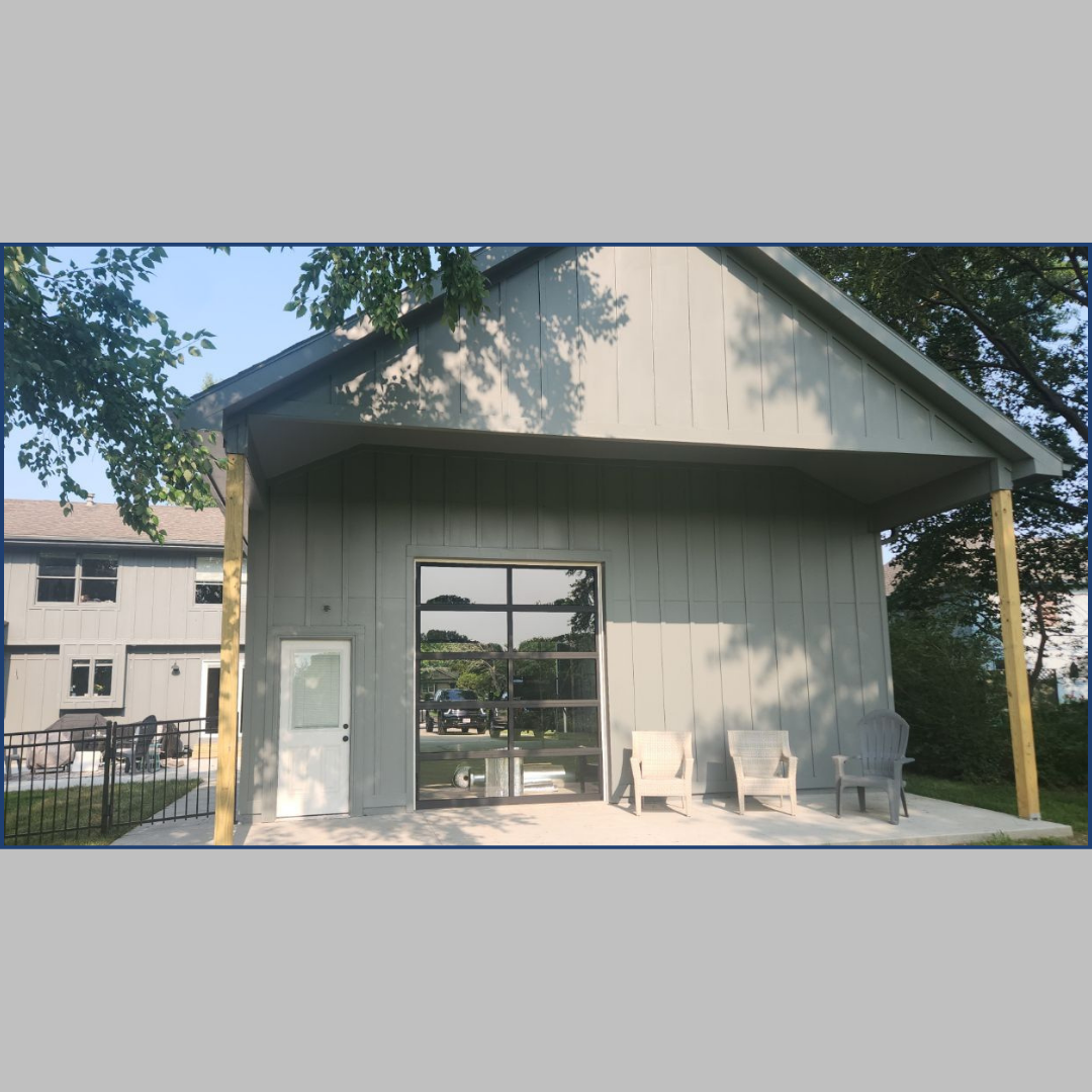 Photo of a modern garage addition with a large glass garage door and a covered patio area. The garage opens to a beautiful lakeside view, with outdoor seating on the patio, offering a blend of functionality and style.