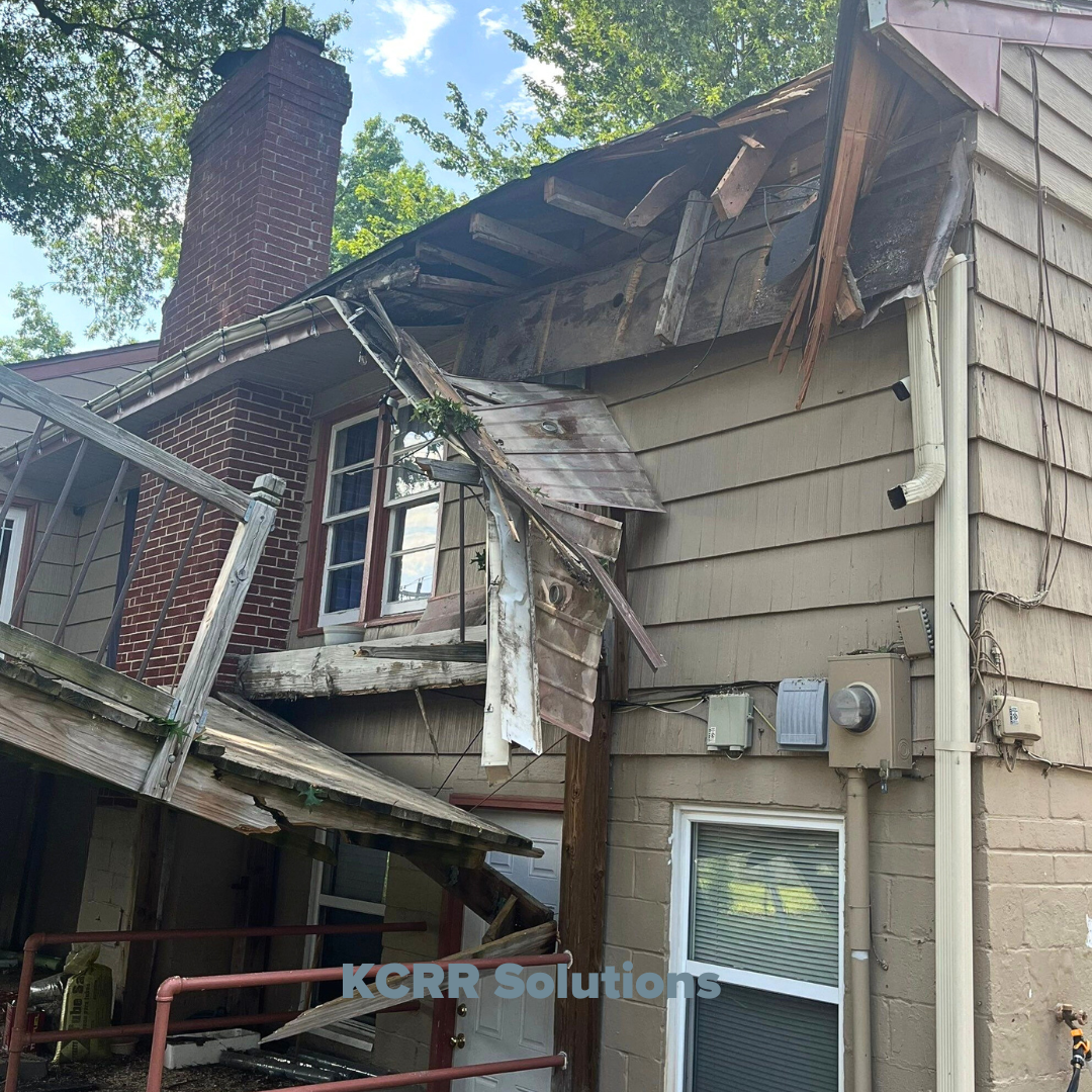 Image showing a severely storm-damaged residential deck knocked off the side of a house, with broken beams, railing, and siding. The impact was caused by a fallen tree, leaving significant structural damage to both the deck and roofline.