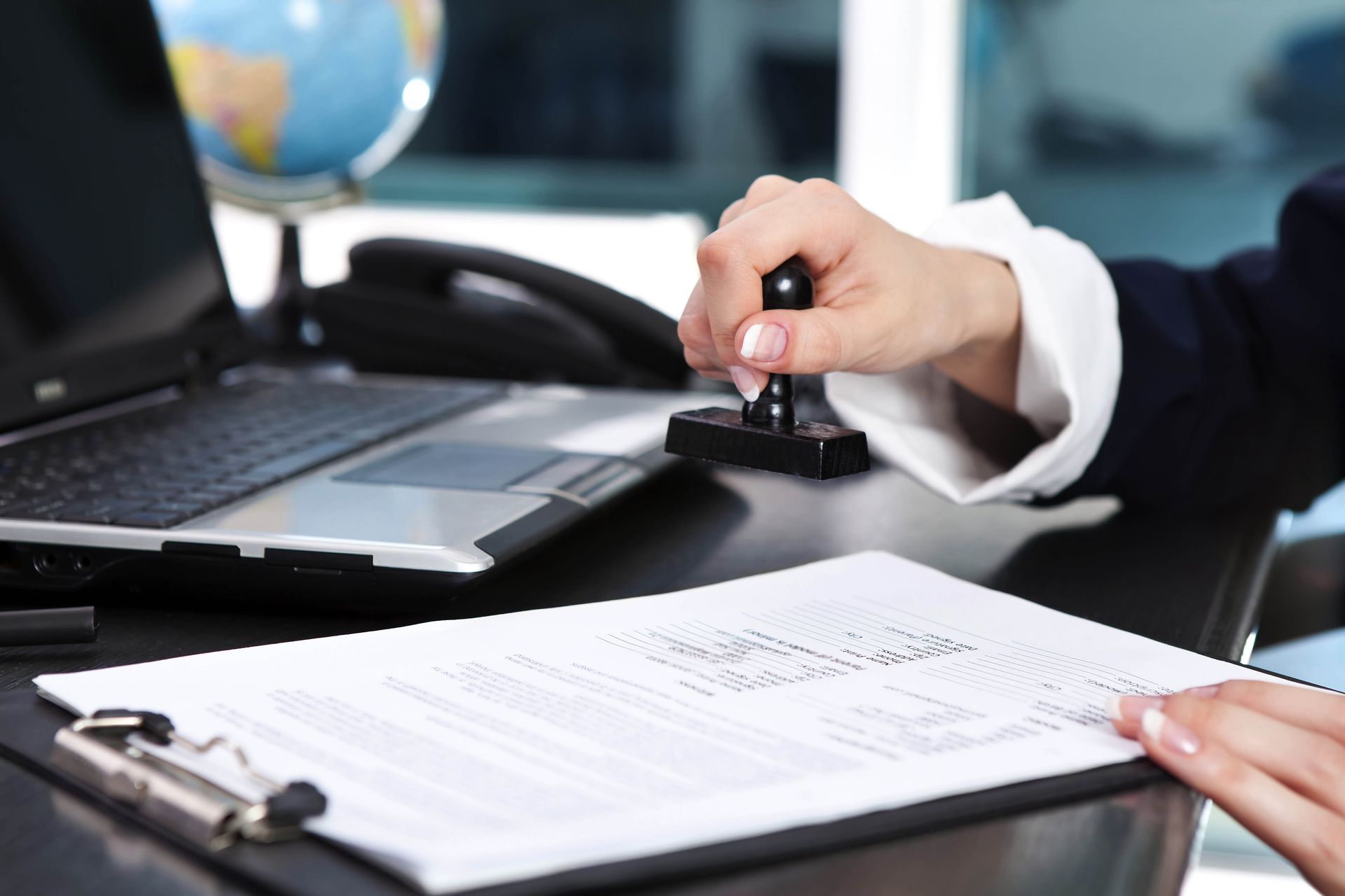 Person stamps document at a desk with a laptop, globe, and phone in office setting.