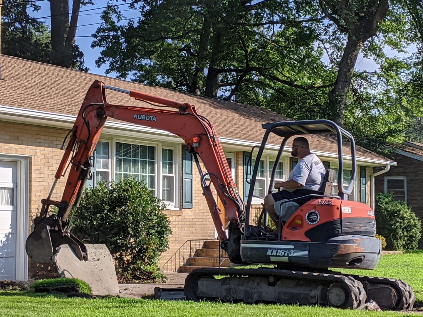 Man operating a small orange excavator in front of a house, digging in the lawn.