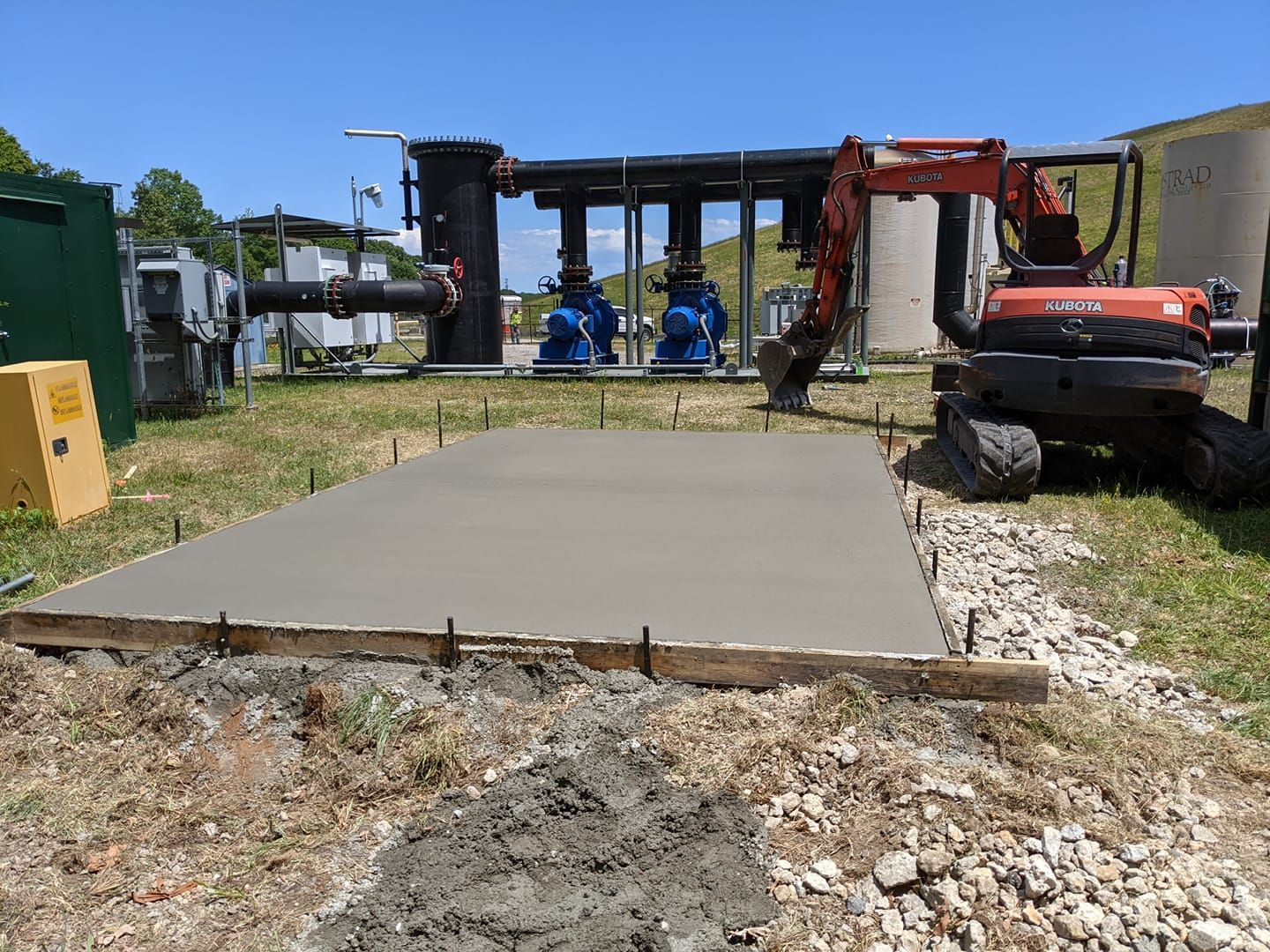Freshly poured concrete pad with wooden forms, excavator, and industrial equipment in the background.