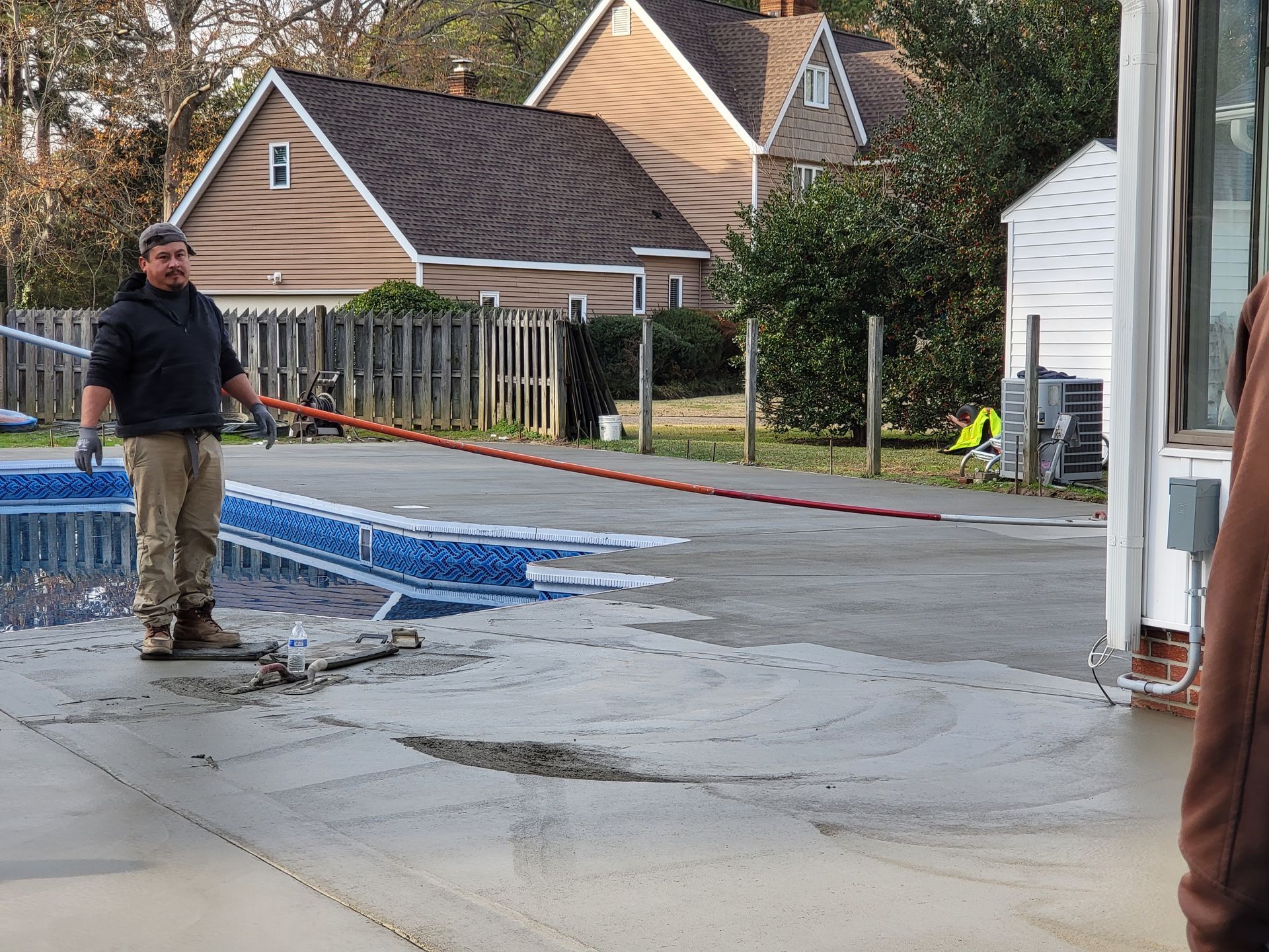 Man smoothing wet concrete on a driveway with a long brush. Houses and a fence are in the background.