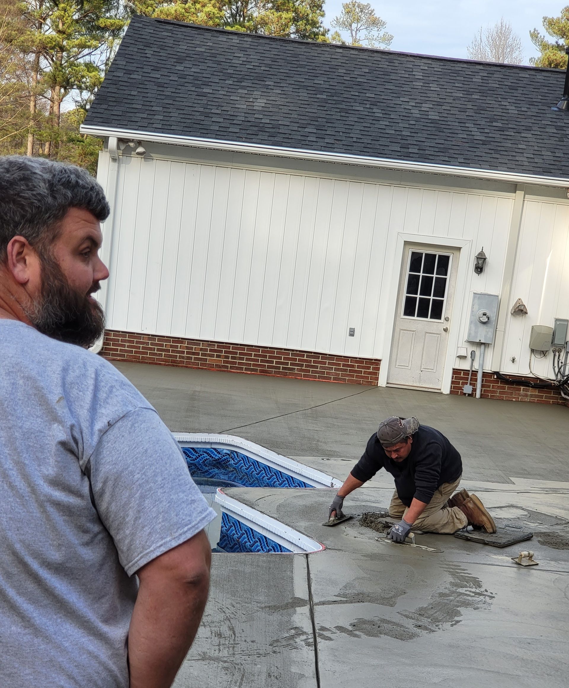 Man watches another working on wet concrete patio by a white building with black roof.