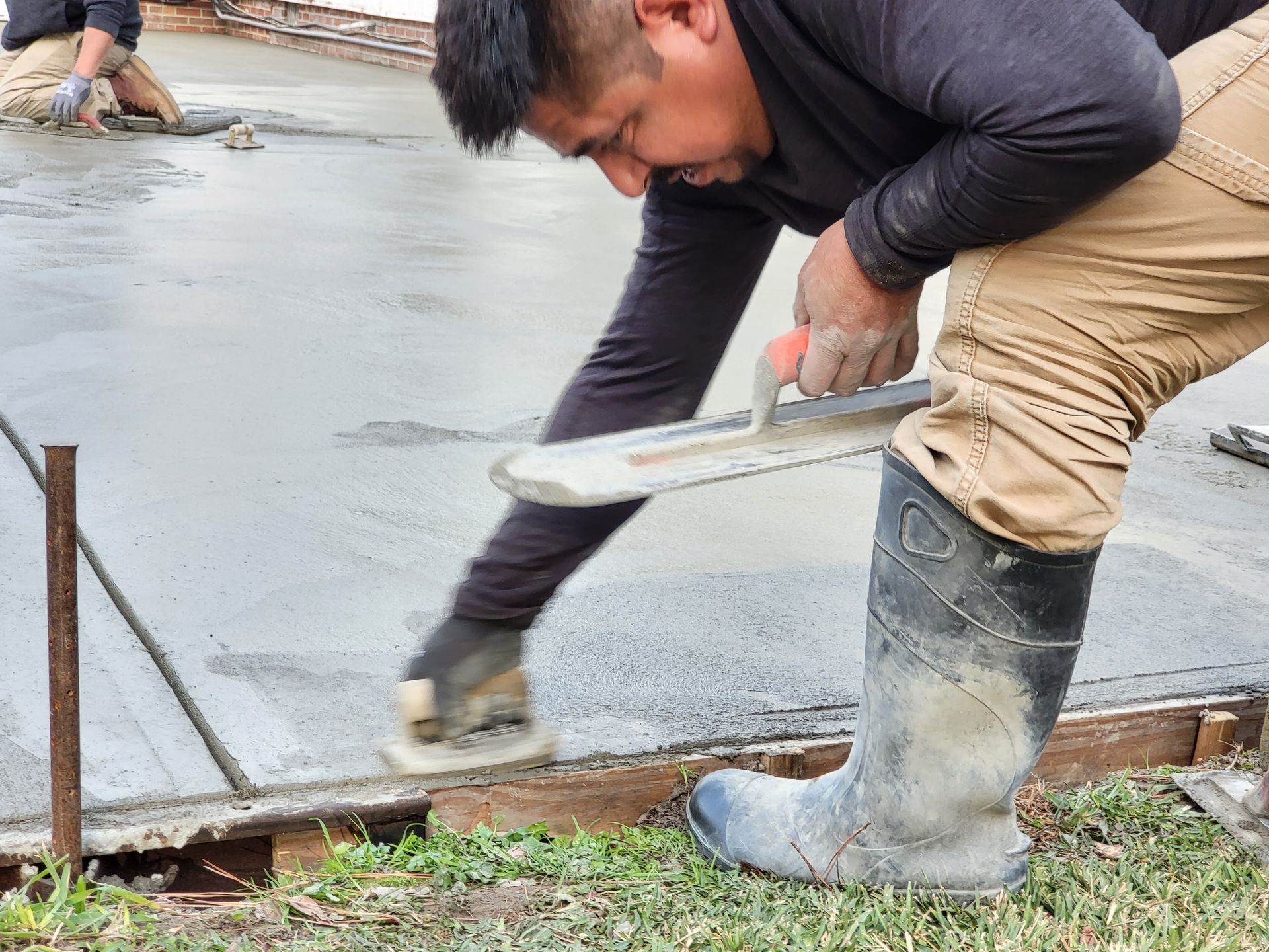 Man smoothing wet concrete with a trowel outdoors, wearing work boots and gloves.