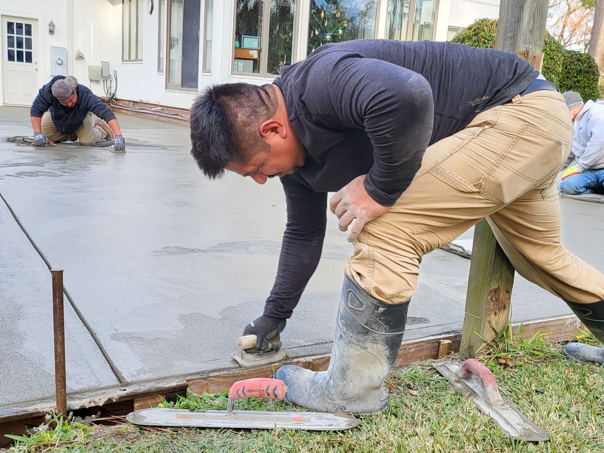 Workers smoothing wet concrete on a driveway. One kneels, hand troweling; others work in background.