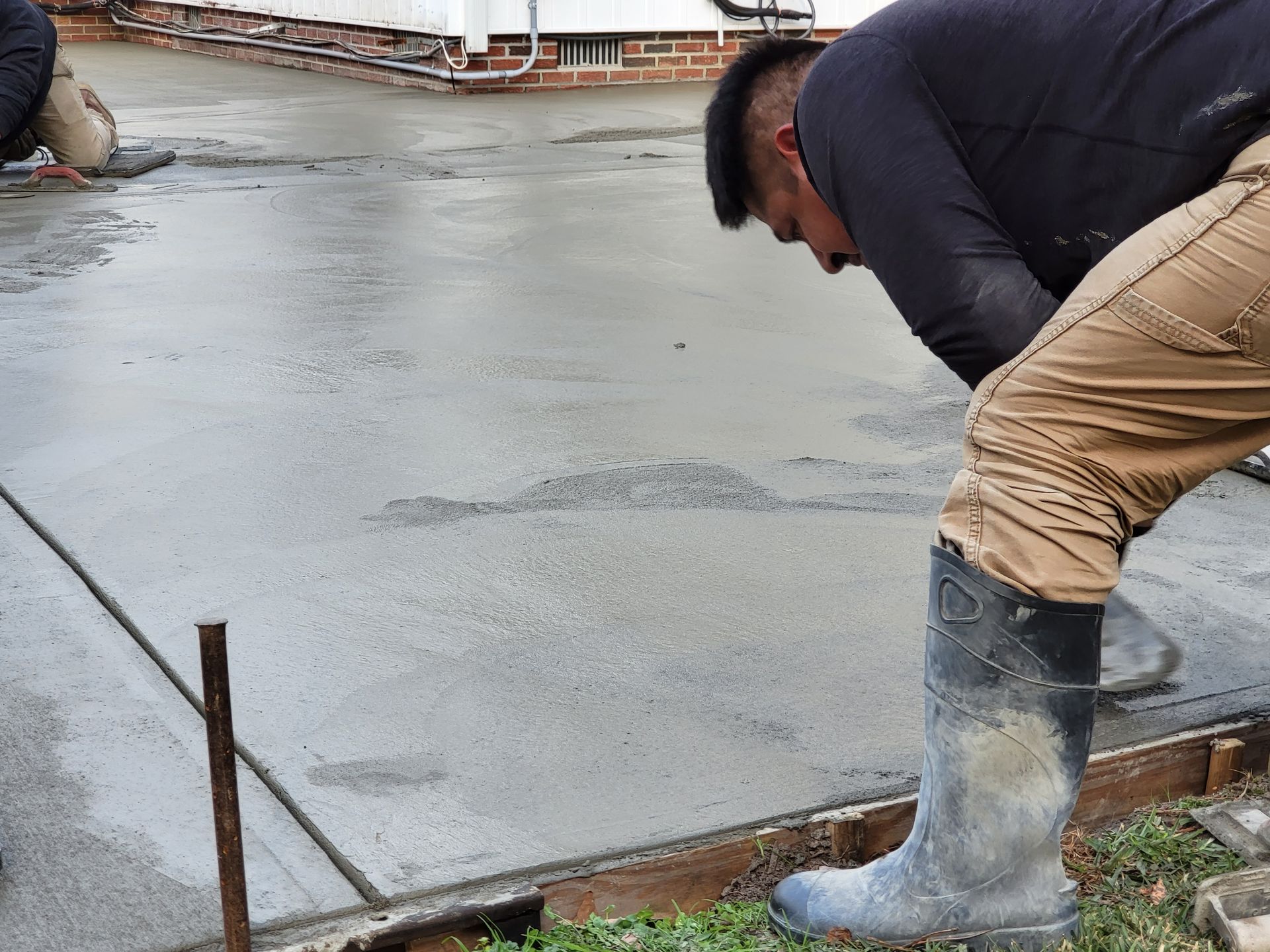 Worker smoothing wet concrete on a sidewalk. Wooden forms, overcast day, grey concrete, dark boots.