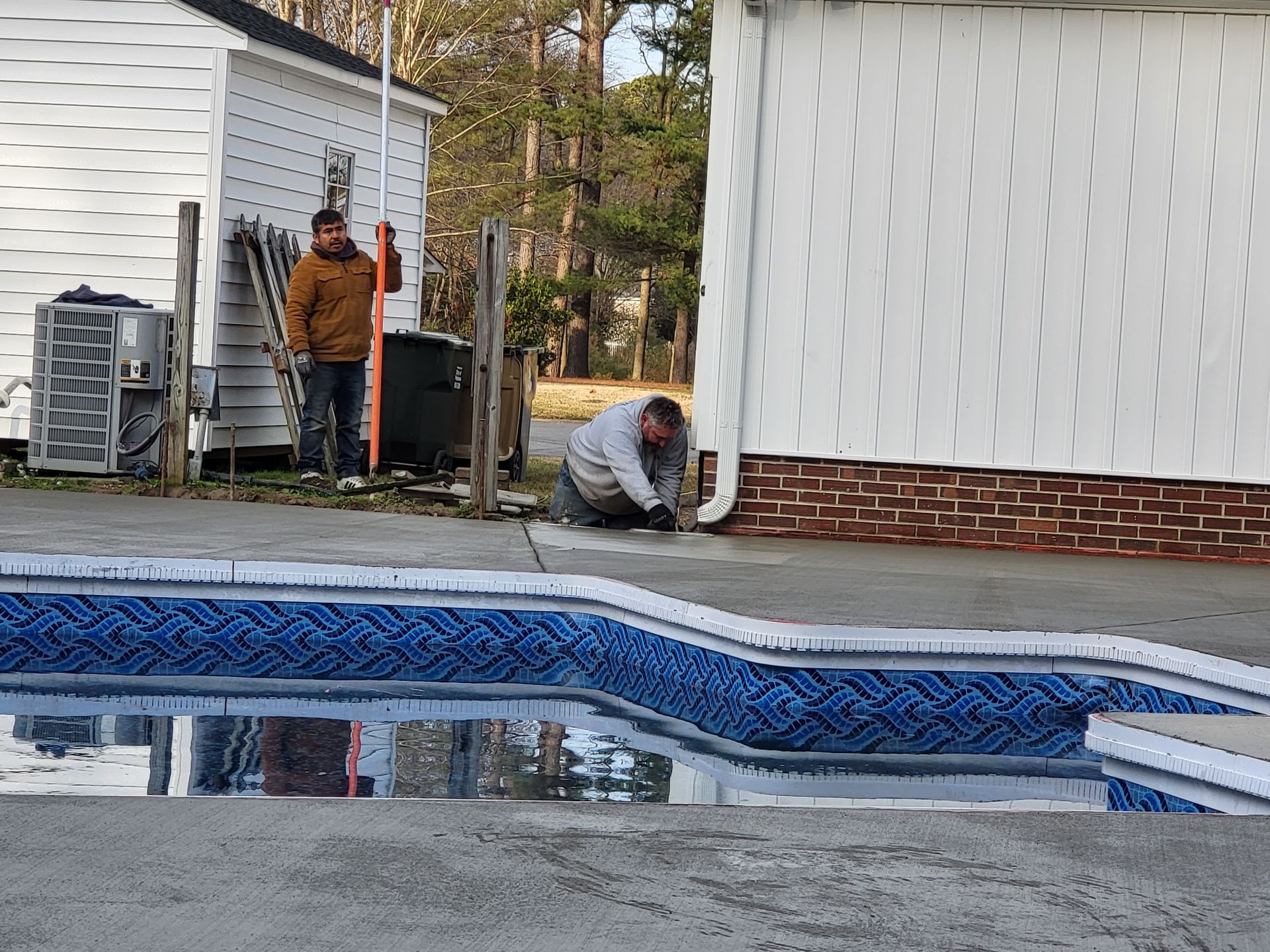 Two men working near a pool. One kneels by building, another stands. White buildings, concrete, and water.