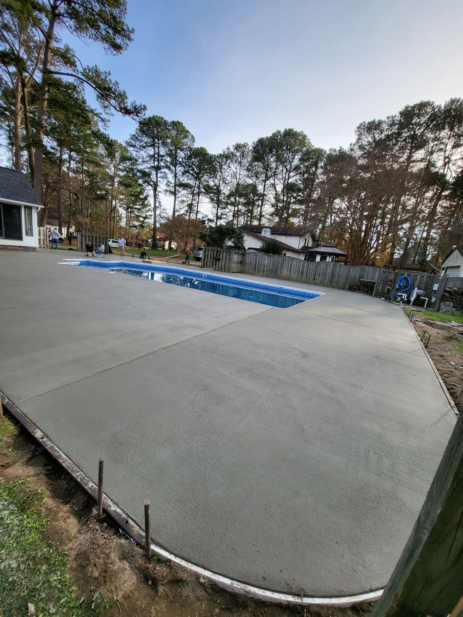 Newly poured concrete patio surrounding a blue-covered swimming pool, in a residential backyard.