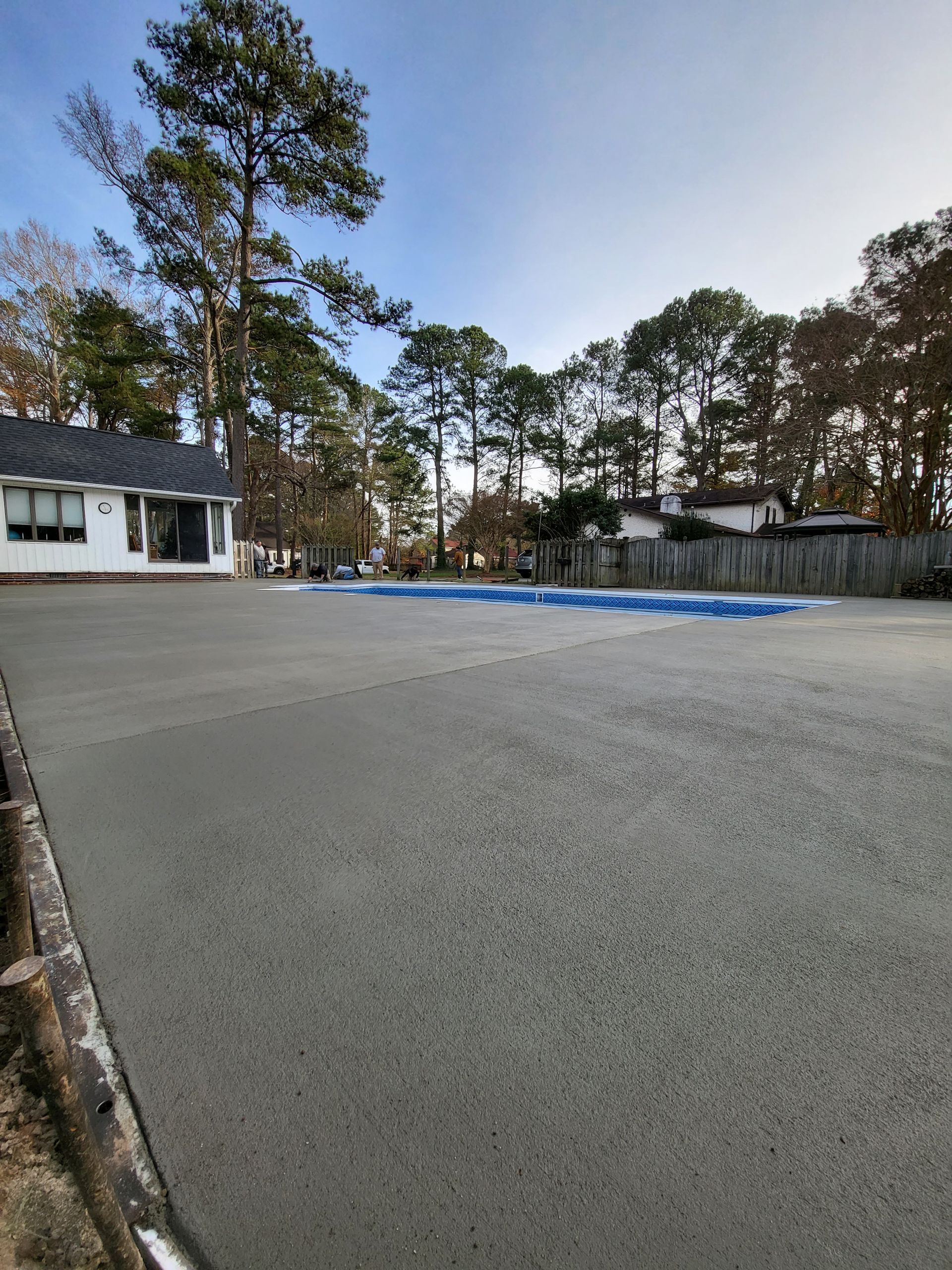 Newly poured concrete patio, a house and trees in the background, blue pool cover visible.