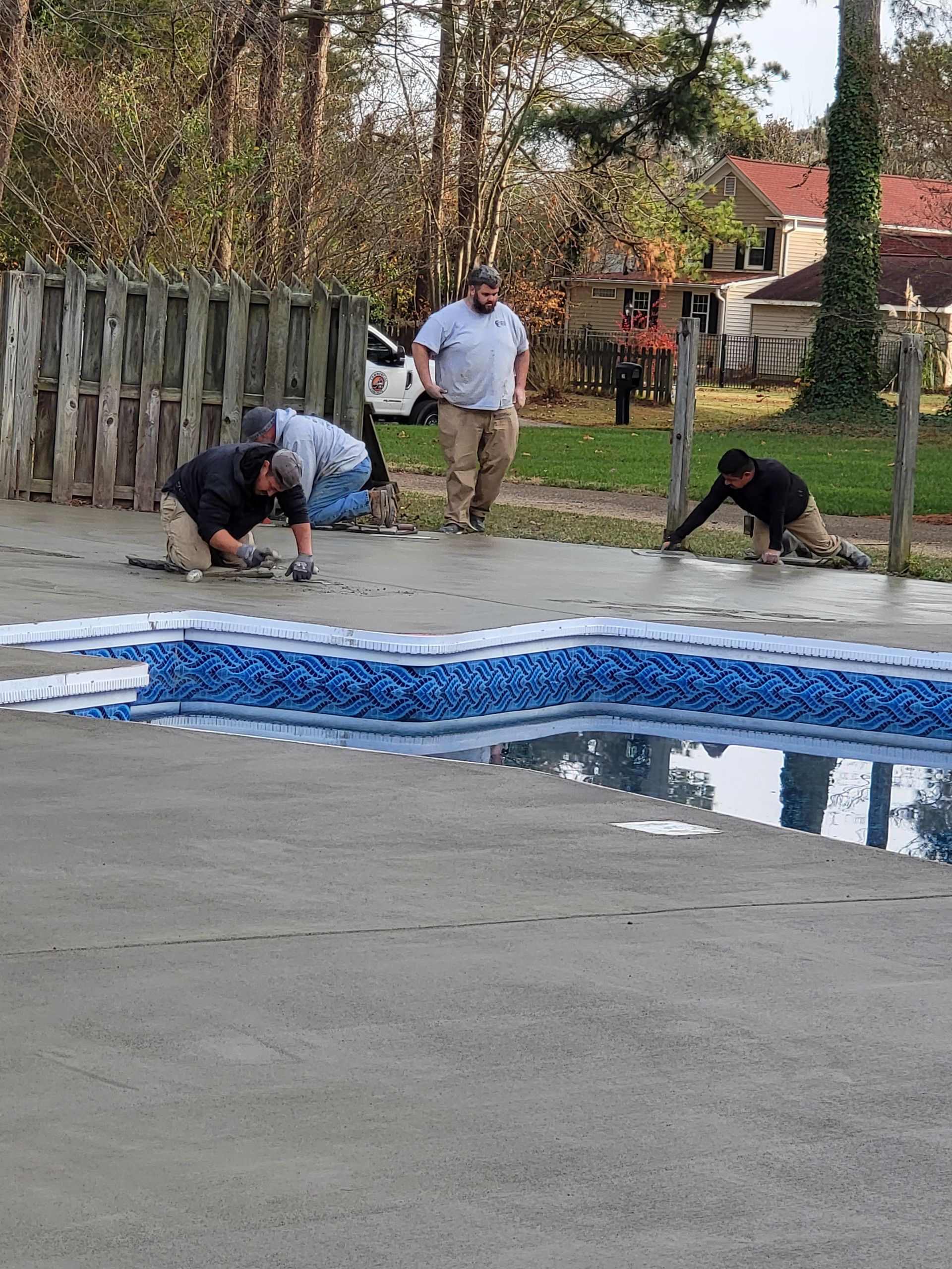 Four people working around a pool, smoothing concrete. Outdoor setting with a fence and trees.