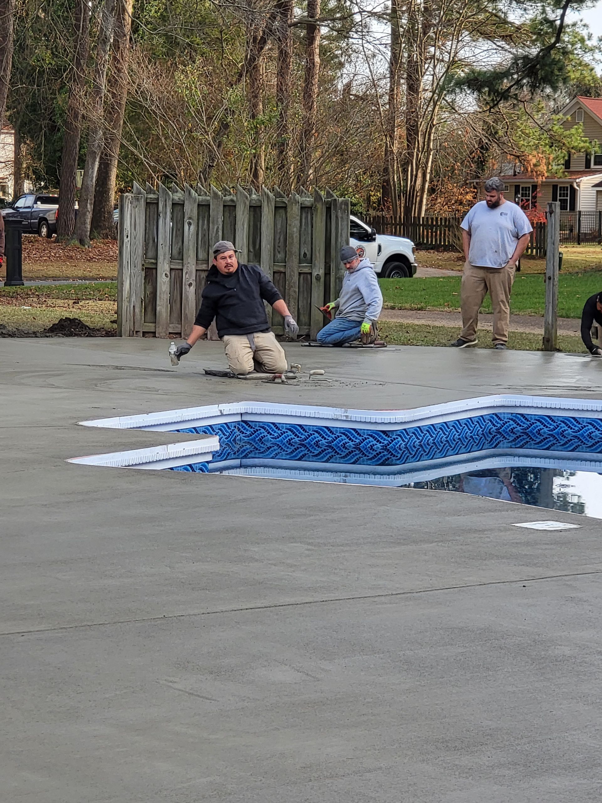 Workers smoothing concrete around a pool. Two are kneeling, another stands, and a truck is visible in the background.