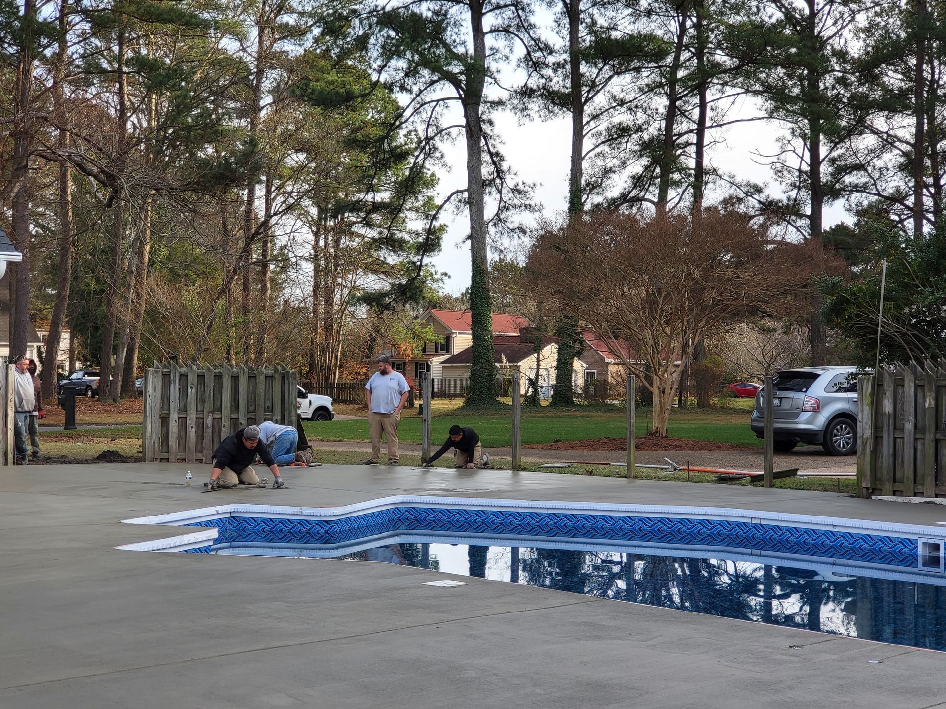 People working on the concrete surrounding a swimming pool in a backyard, with a car and trees in the background.