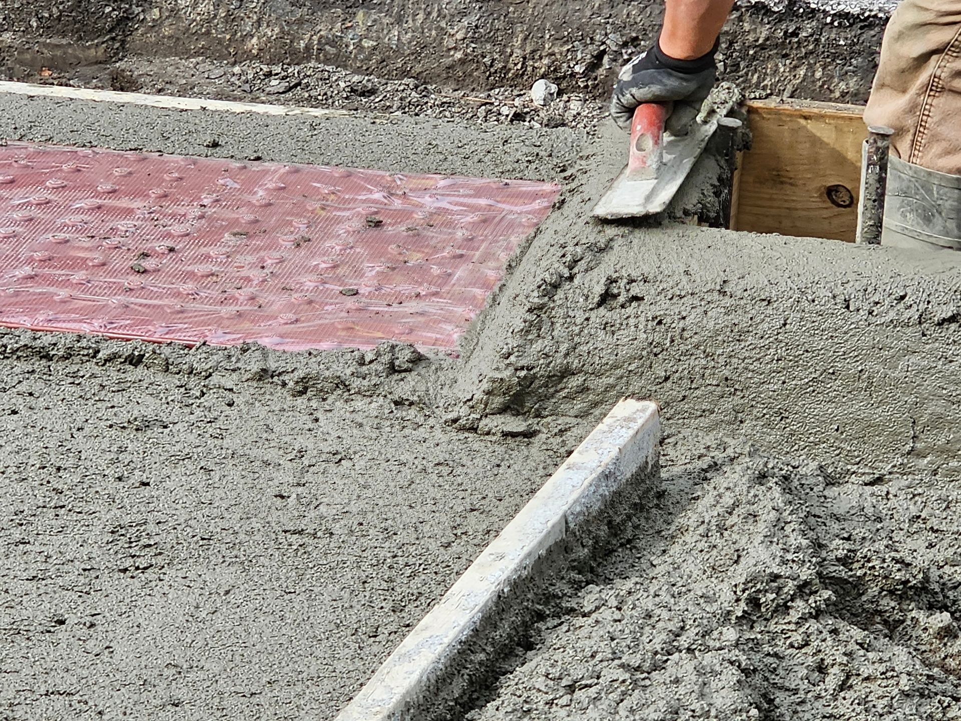 Person smoothing wet concrete with a trowel, next to a red-tinted concrete section.