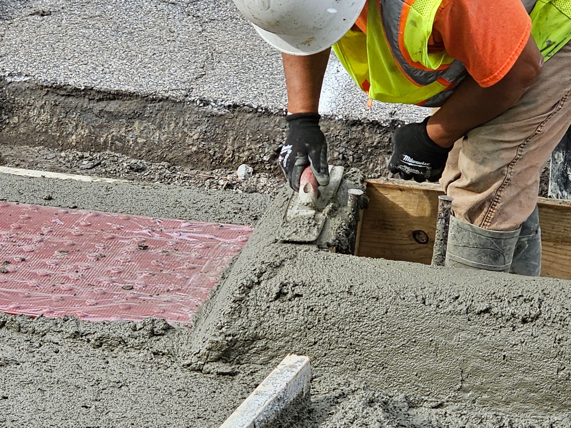 Construction worker smoothing concrete with a trowel, wearing safety vest and gloves.