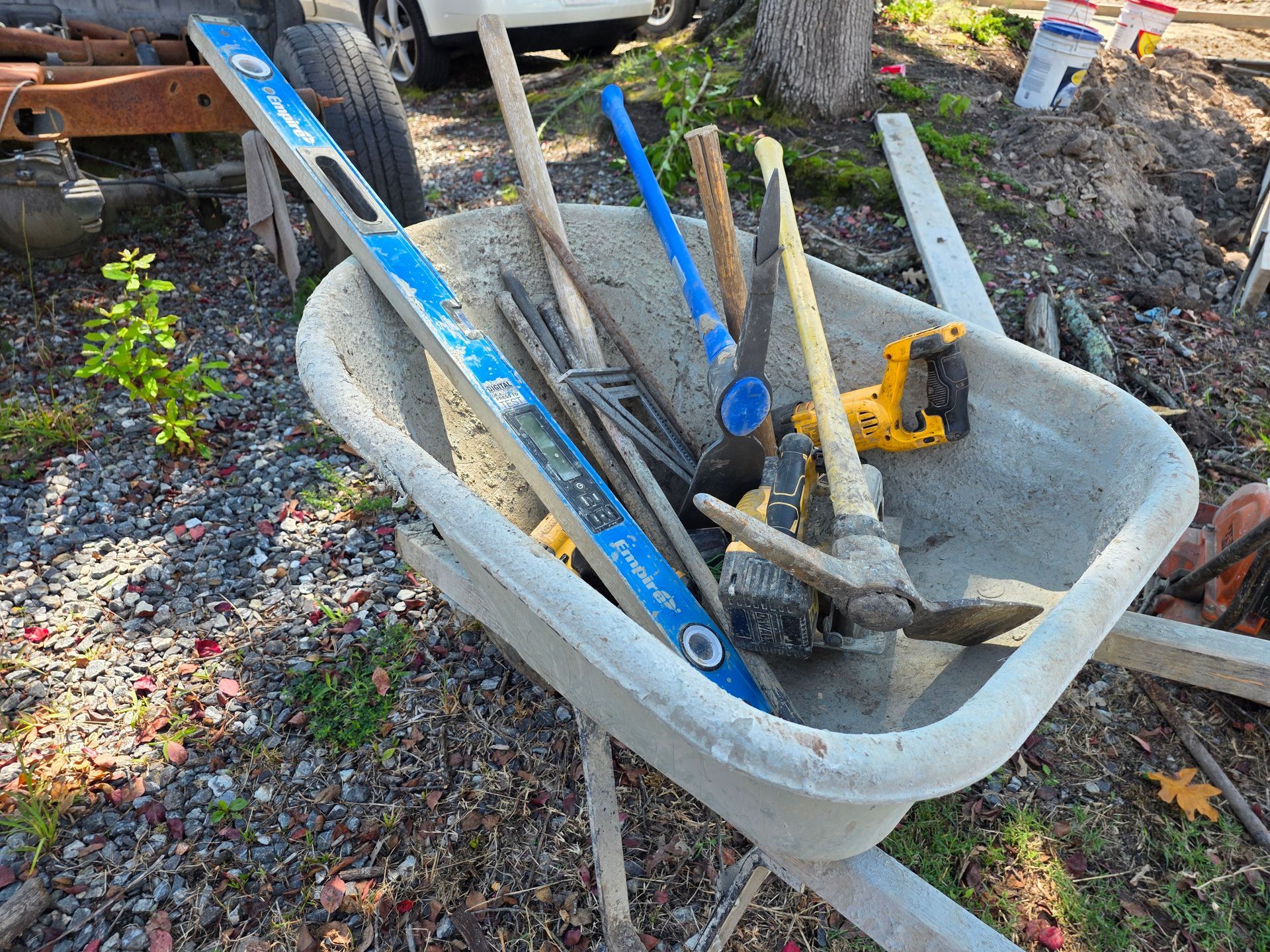 Wheelbarrow filled with tools, including a level, pickaxes, and a saw, outdoors.