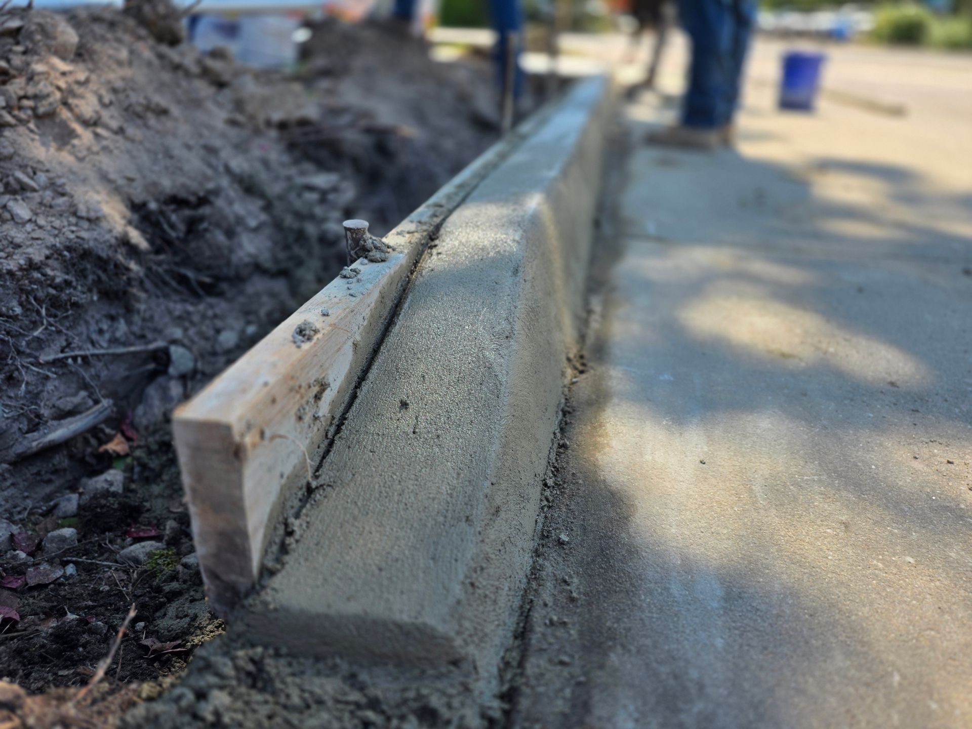 Freshly poured concrete curb with wooden formwork next to dirt and a paved road; construction.