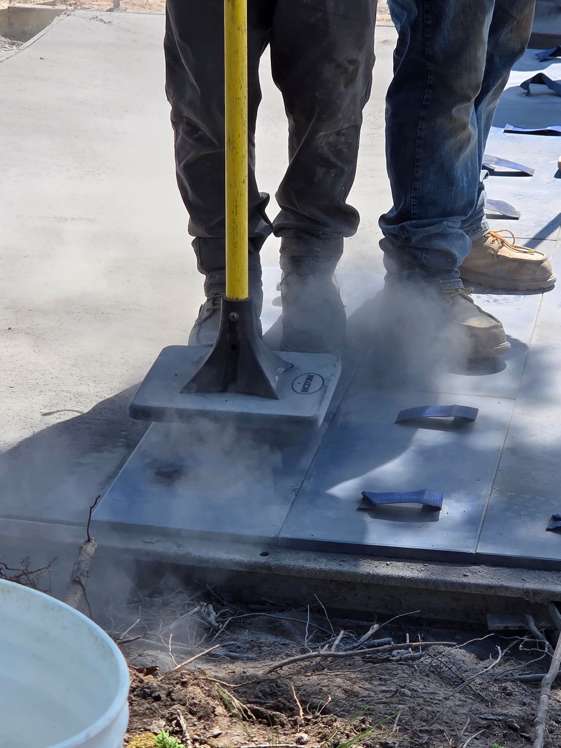 Workers using a hand tamper on a concrete surface, dust rising.