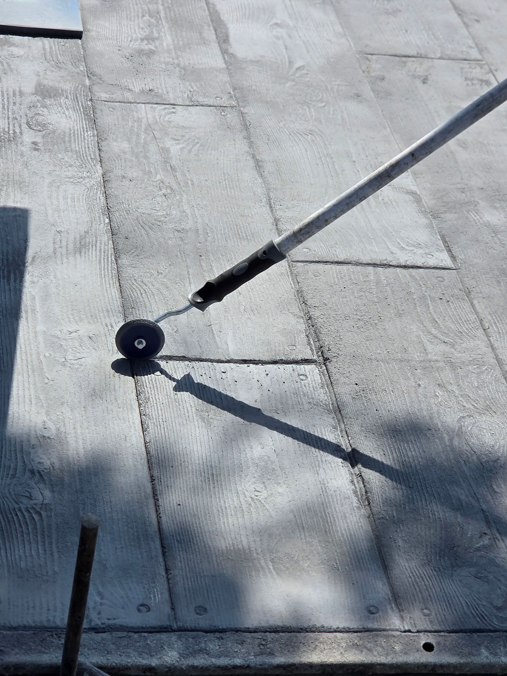 A person uses a roller on a flat roof. The roller is gray, and the roof is made of rectangular gray panels.
