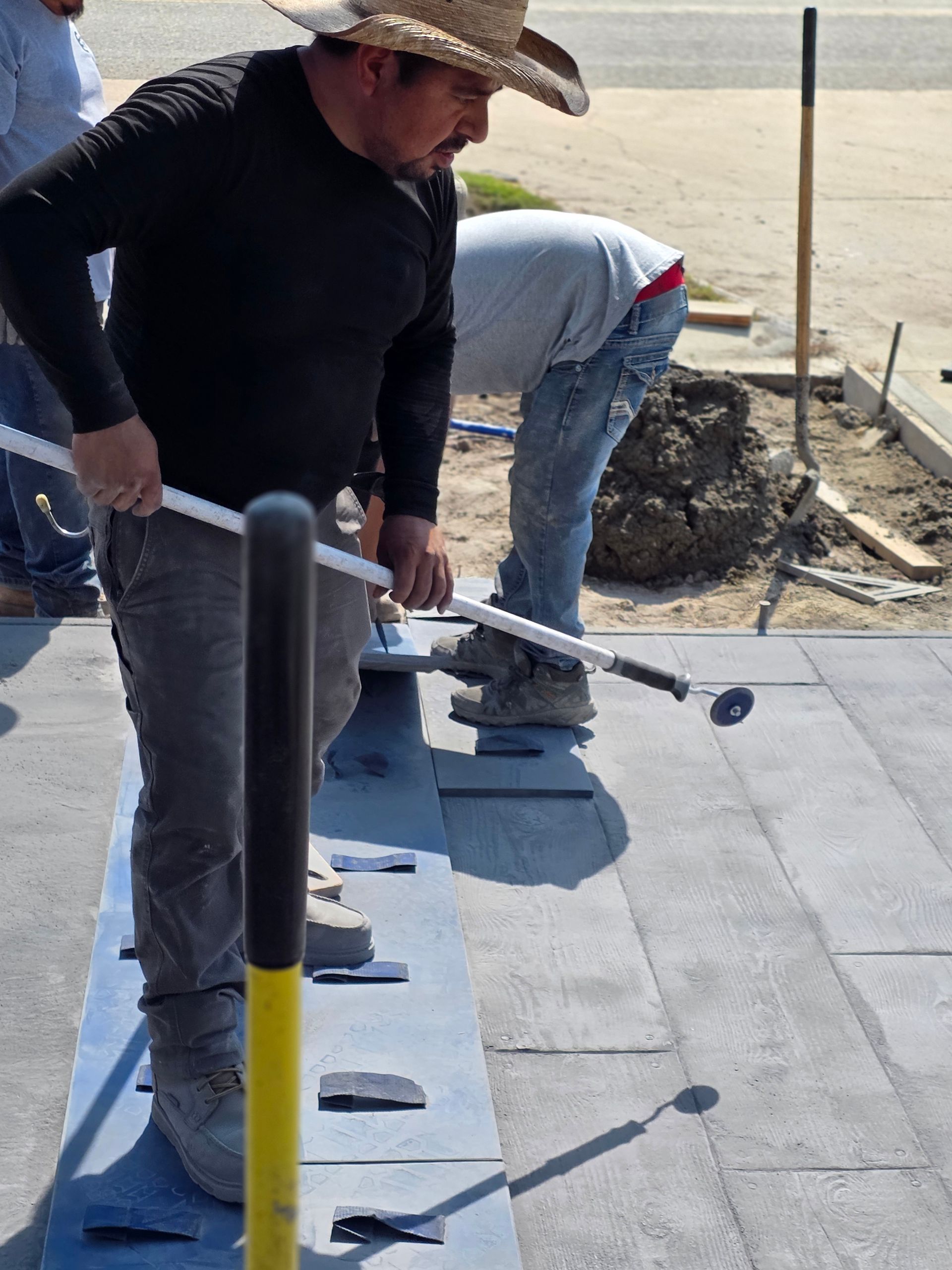 Construction worker using a long-handled tool to work on a concrete sidewalk. Another worker visible nearby.