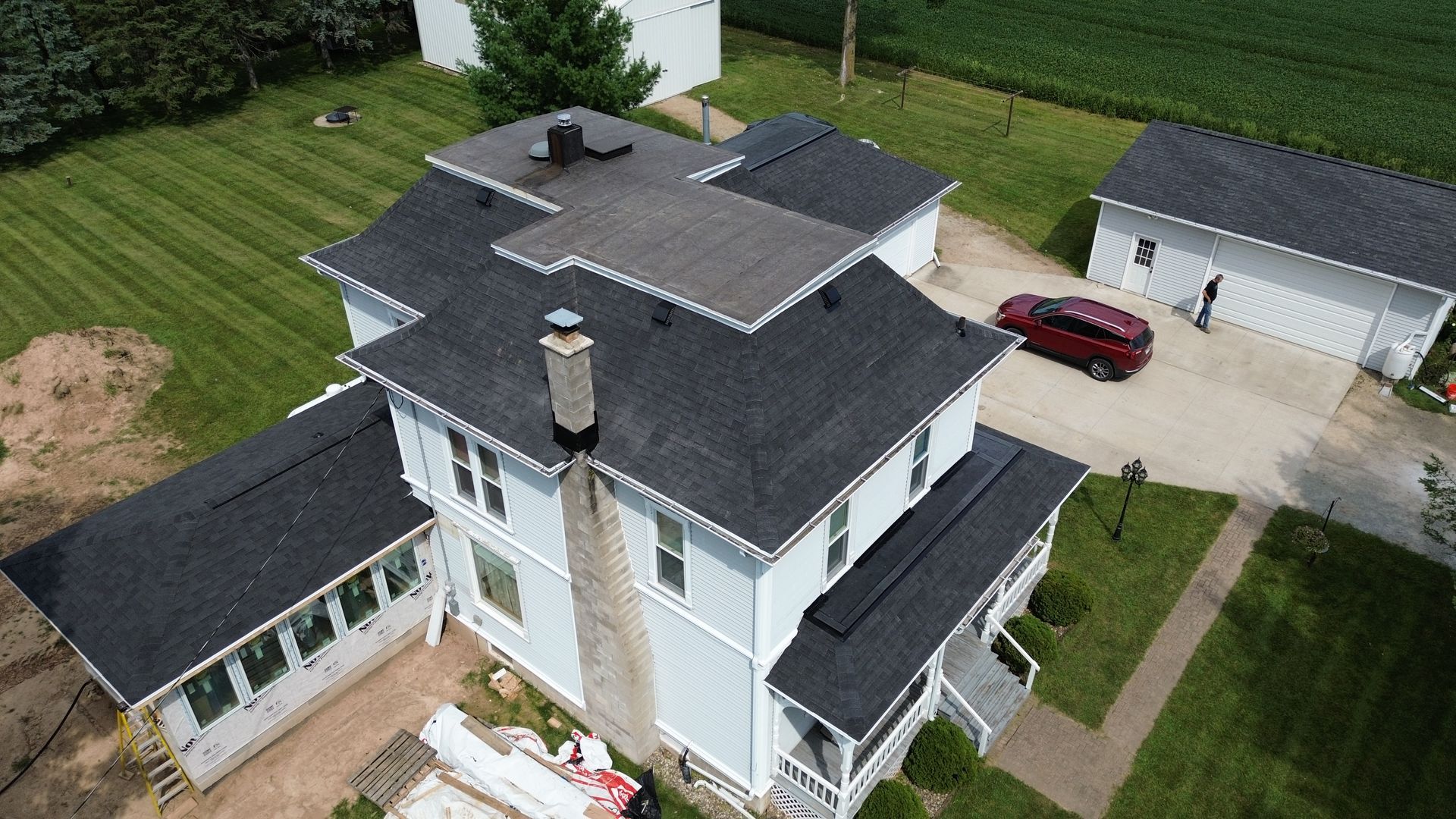 Aerial view of a light blue house with a dark gray roof, chimney, and attached garage, red car parked on gravel.