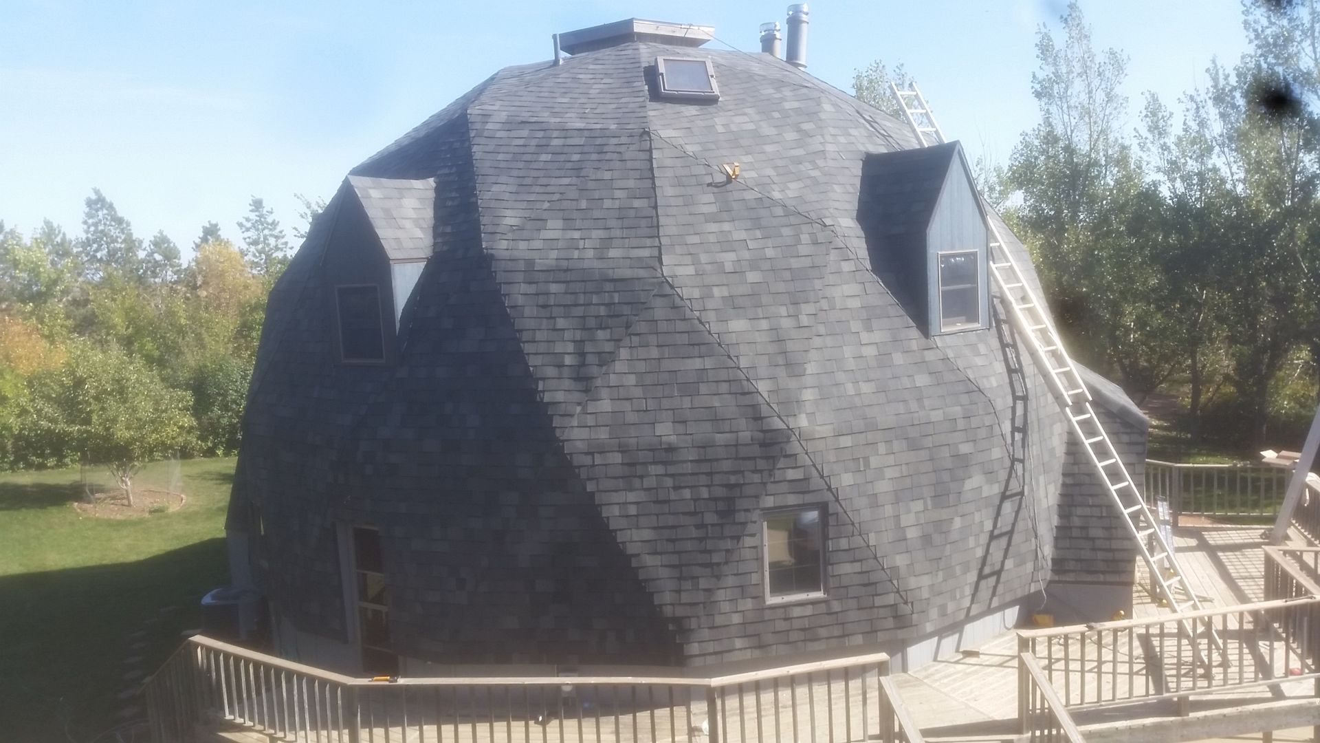 Geodesic dome house with gray shingles and a wooden deck on a sunny day.