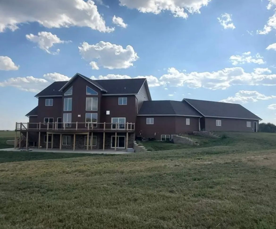 Large brown house with a deck, set on a grassy hill under a blue sky with white clouds.