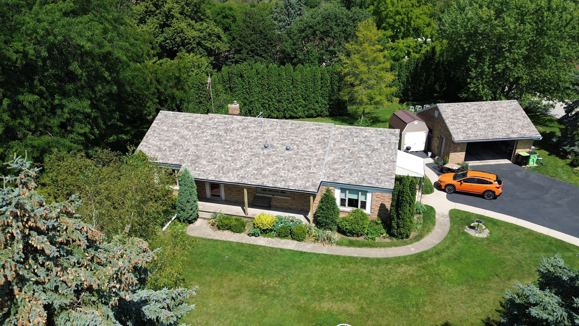 Aerial view of a ranch house with a garage and orange car on a green lawn, surrounded by trees.