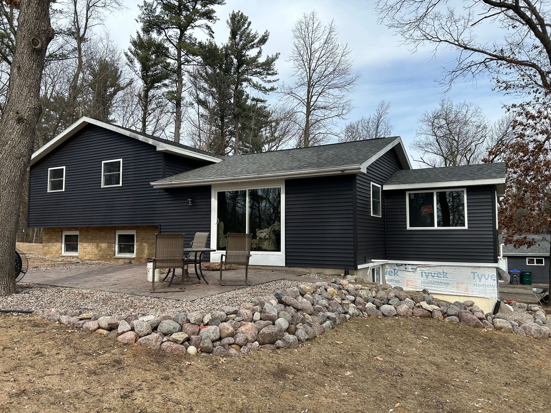 Dark gray house with white trim, a small patio, and a rock retaining wall.