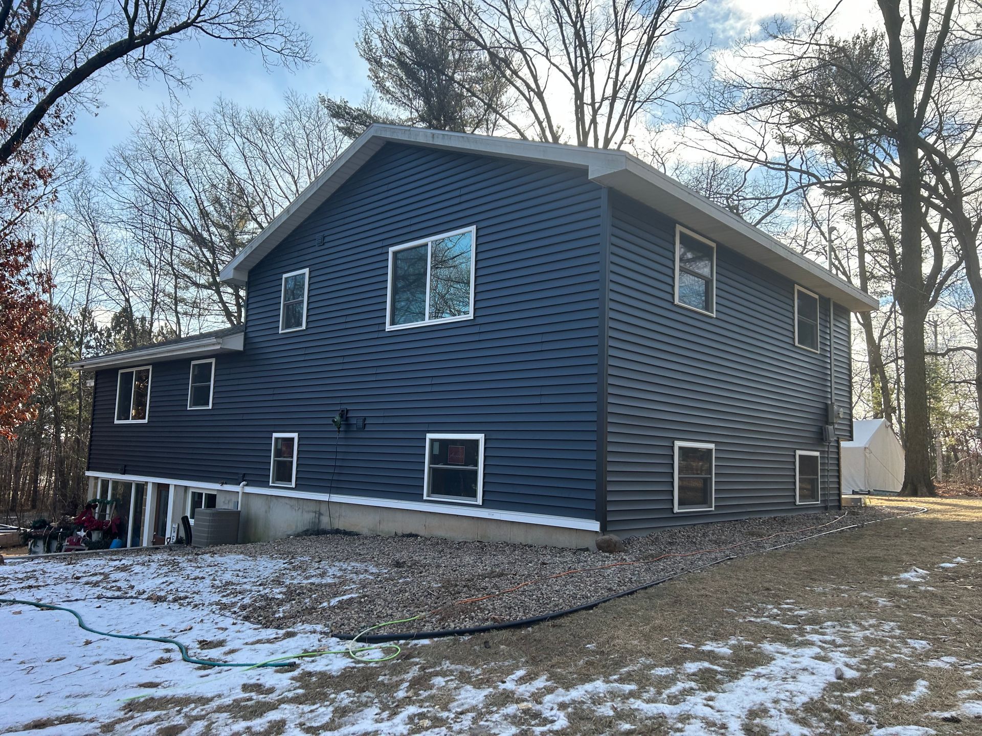 Two-story navy blue house on a snowy, wooded lot with multiple windows, bright sky.