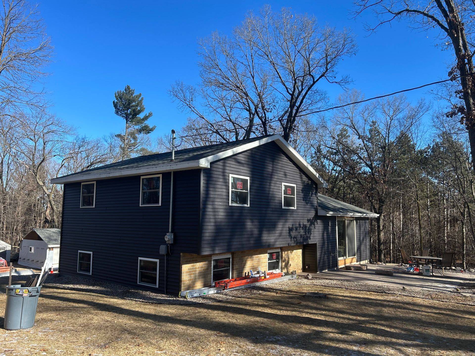 Two-story house with dark blue siding, tan foundation, and bare trees against a blue sky.