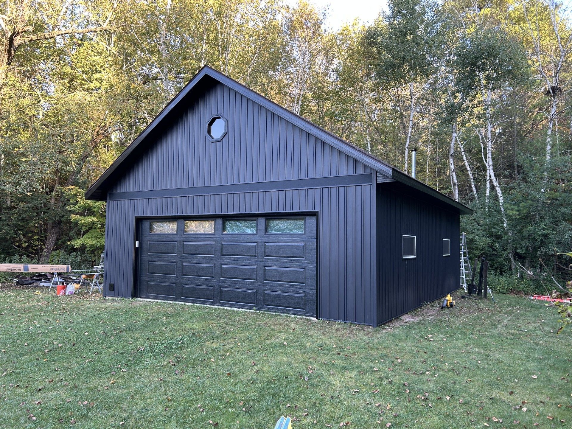 Black garage with a round window, set on a grassy yard with trees in the background.