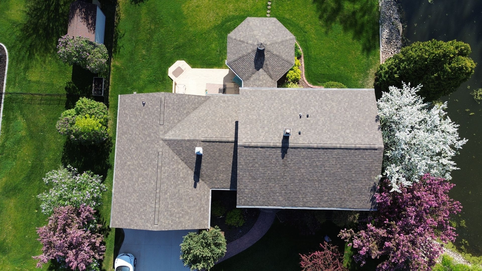 Overhead view of a house with a gazebo, driveway, surrounded by green grass, trees, and water.