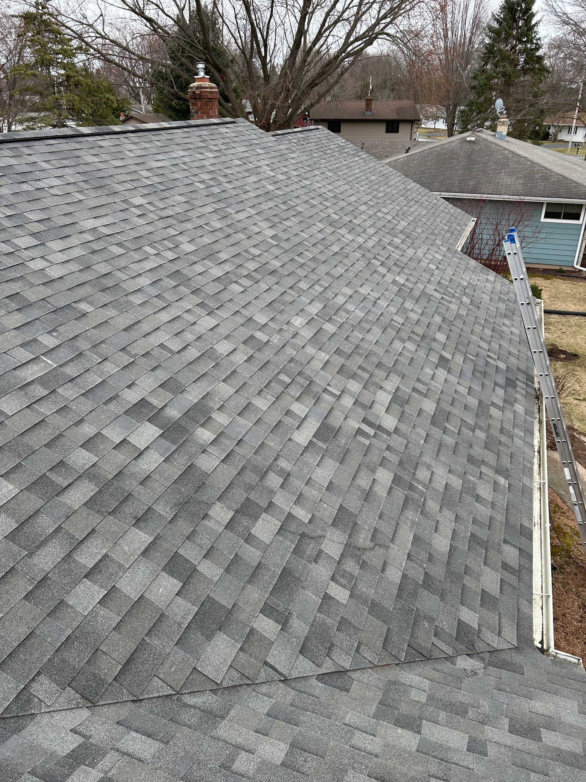 Gray asphalt shingle roof on a house. Chimney and trees in the background. Cloudy day.