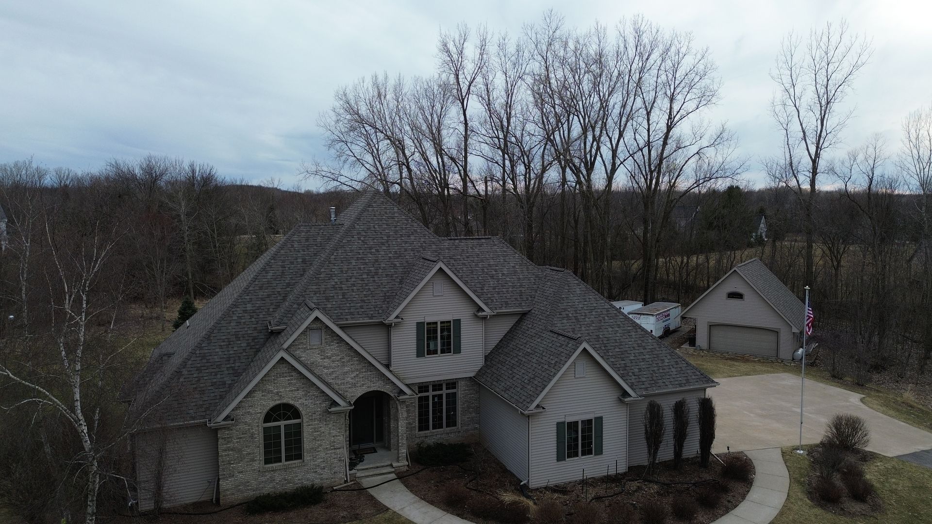 A large, two-story house with a stone facade sits amidst bare trees under a cloudy sky.