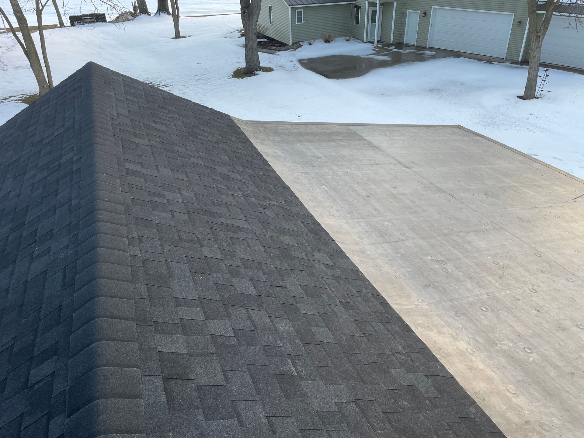 Dark shingled roof next to a light-colored flat surface, possibly a driveway, with snow in background.