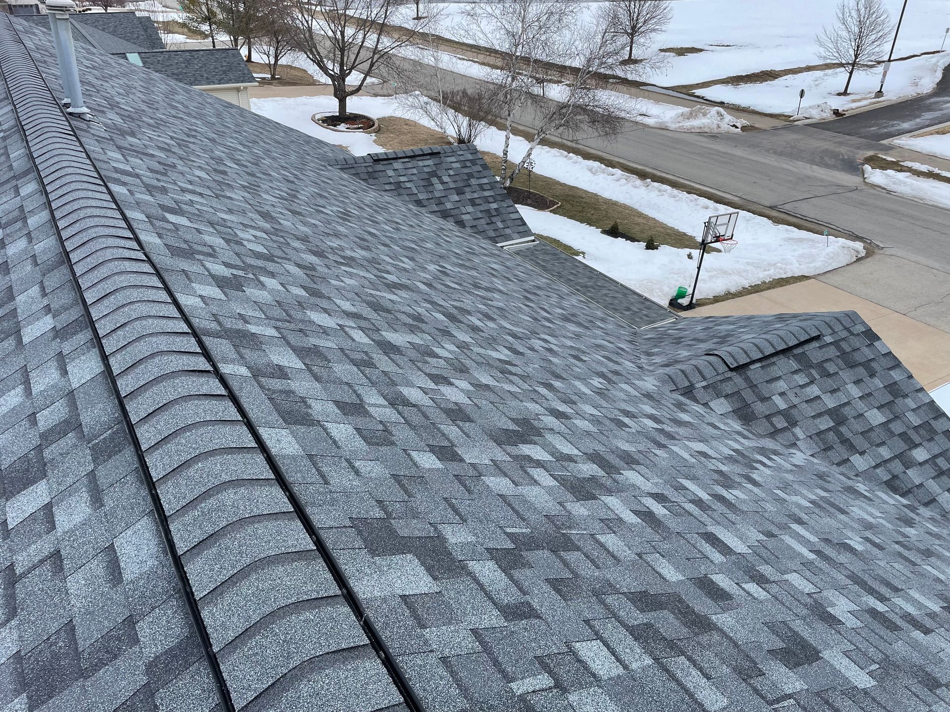 Gray asphalt shingle roof on a house, with snow on the ground and trees visible in the distance.