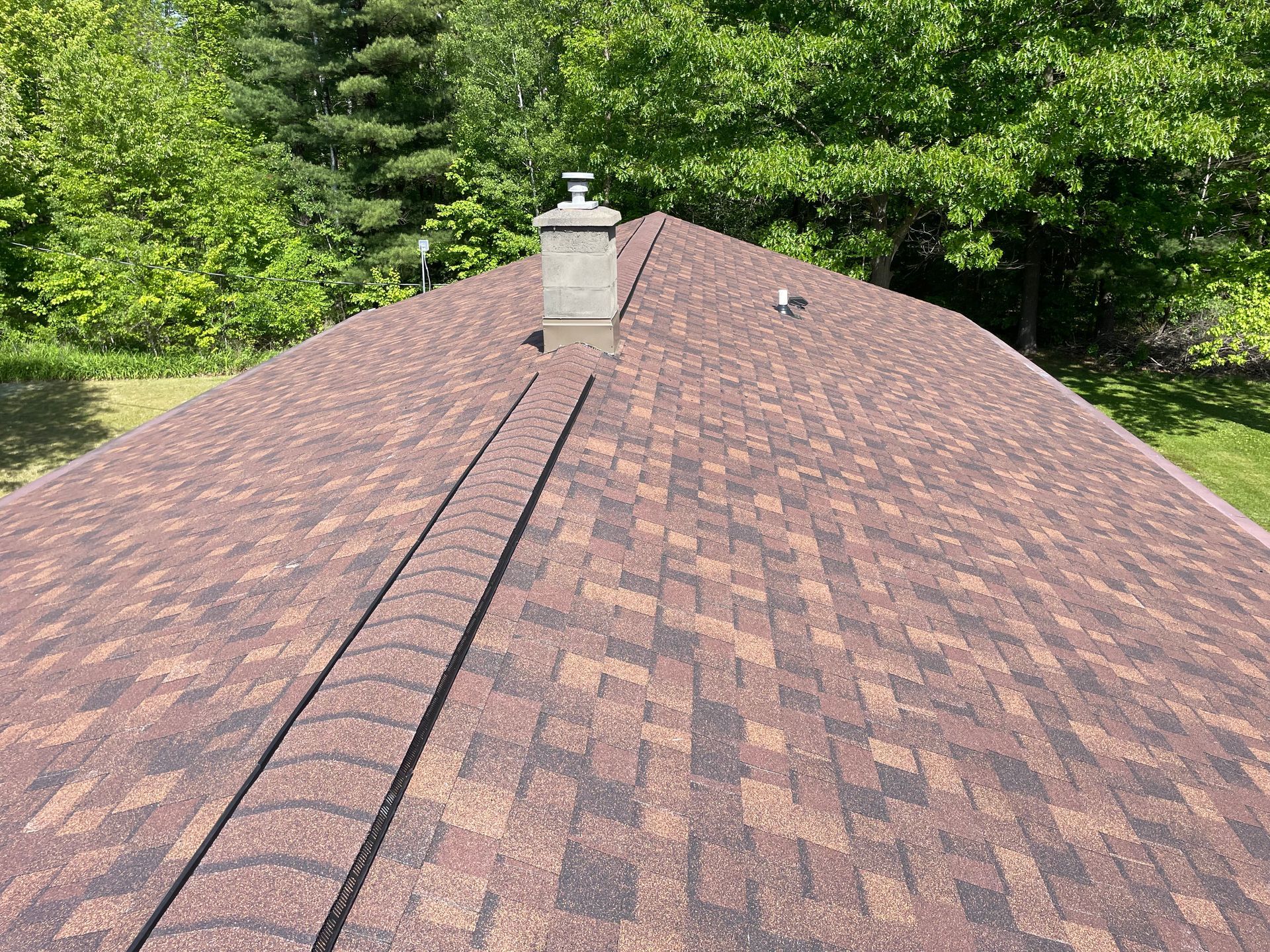 Brown shingle roof with a chimney, trees in the background on a sunny day.