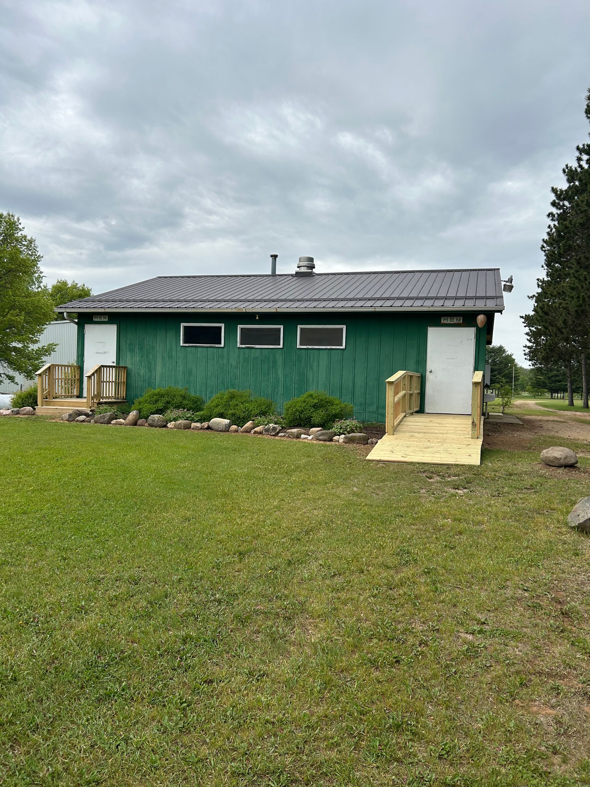 Green building with white doors, windows, and a ramp for accessibility on a grassy lawn under a cloudy sky.