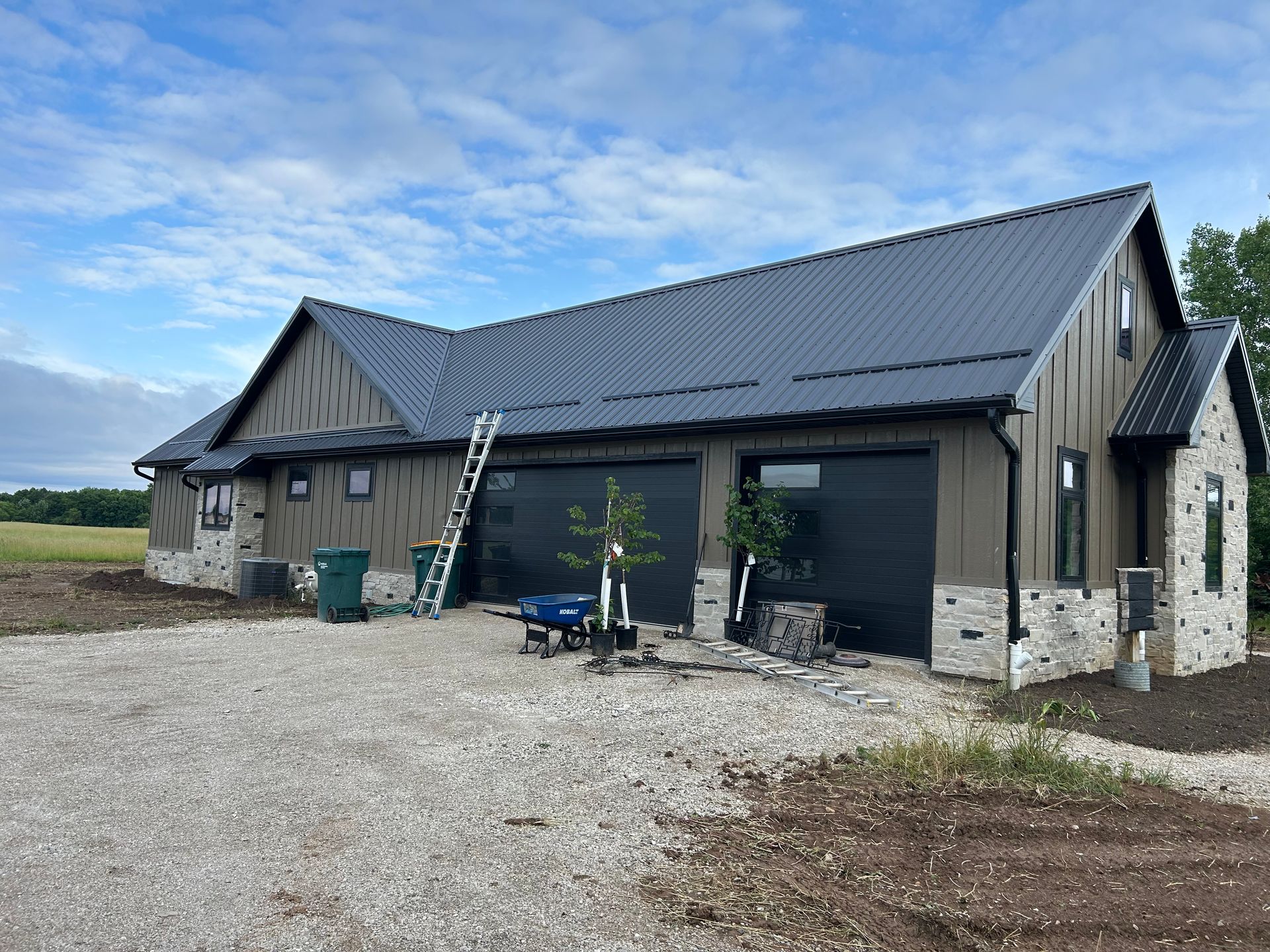 Modern home with dark metal roof and garage doors, tan siding, and stone accents. Gravel driveway and sky.